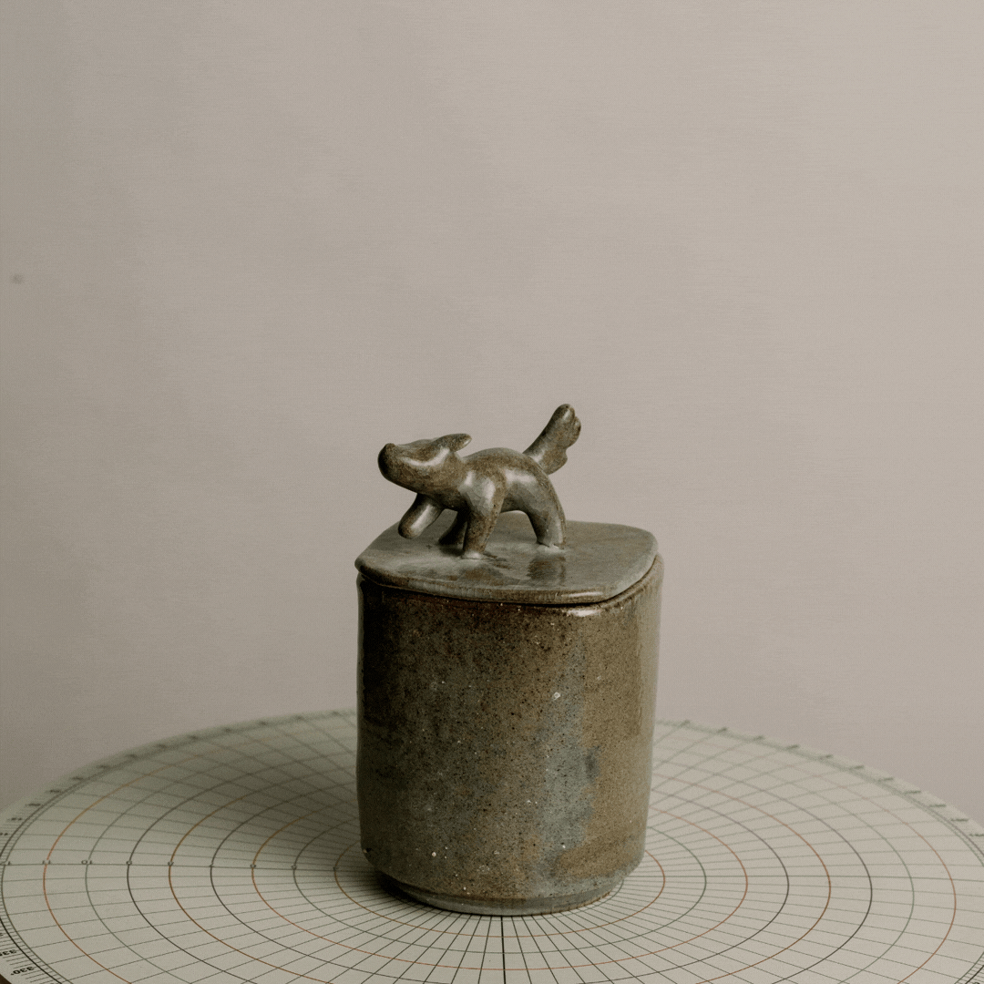 Ceramic container with a small abstract animal sculpture on top, placed on a circular grid-patterned surface.