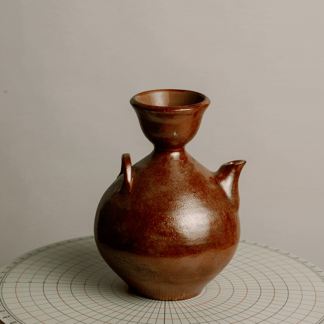 A brown ceramic vessel with a rounded body, a narrow neck, a flared rim, and two small spouts on the sides, placed on a circular grid surface.