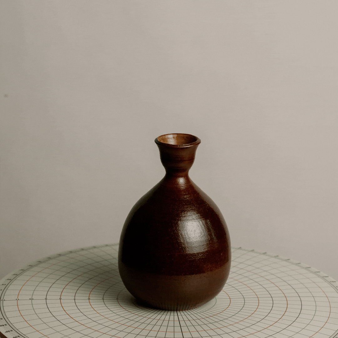 A small, brown ceramic vase with a glossy finish on a round measurement chart with concentric circles, against a plain light-colored background.