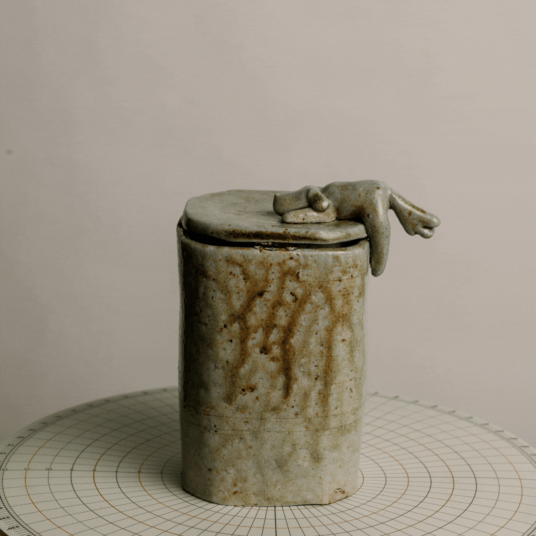 A rustic, cylindrical ceramic container with a lid, featuring a small, irregularly shaped ceramic hand resting on top. The container has a weathered, glazed surface with brownish stains and is placed on a grid-lined surface against a plain background.