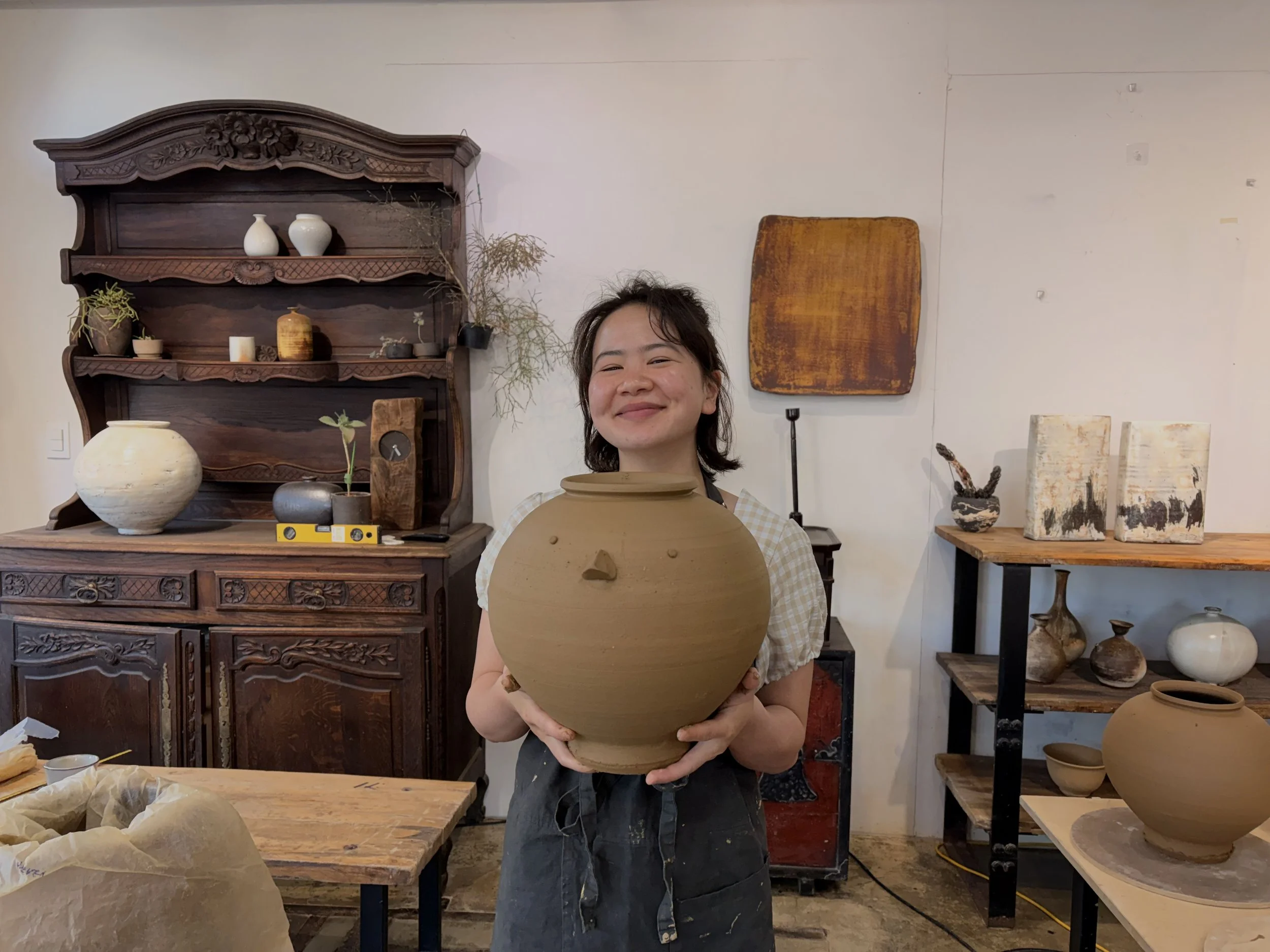 A woman standing in a pottery studio holding a large, round, unglazed clay vase. She is smiling, wearing a checkered apron, with various pottery and decorative objects on shelves behind her.