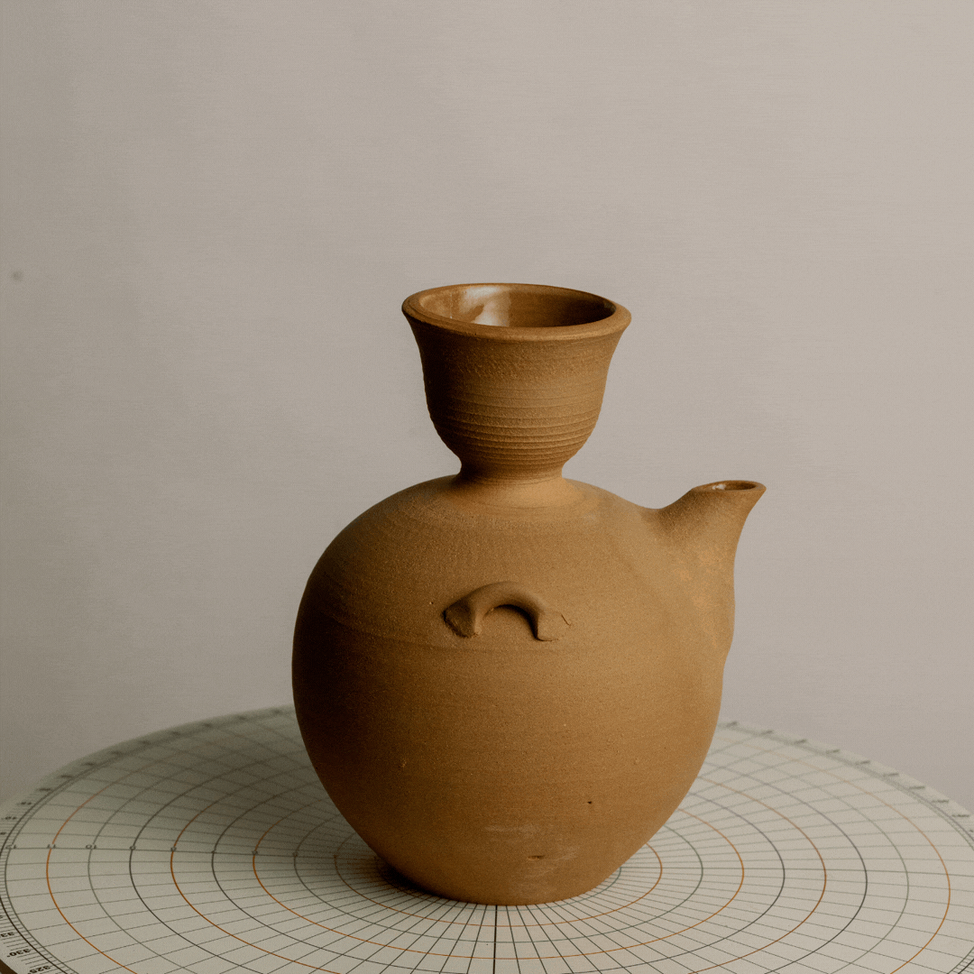 Clay pottery with a round body, small spout, and an upside-down cup on top, placed on a grid-patterned surface.