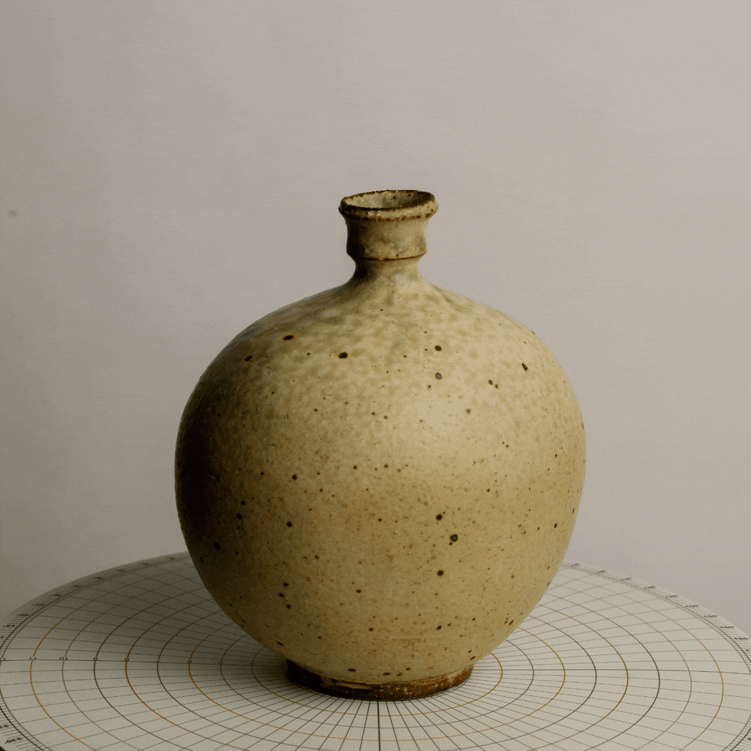 A beige, speckled ceramic vase with a small opening at the top, placed on a circular grid surface with measurement markings.