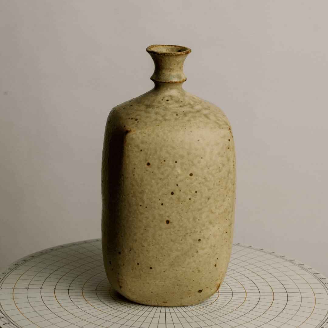 A beige ceramic bottle with a narrow neck and wider base, placed on a circular grid-patterned surface against a plain background.