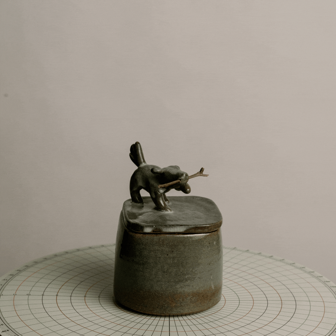 Small ceramic container with a lid, topped by a figurine of a dog holding a stick, on a grid-lined surface.