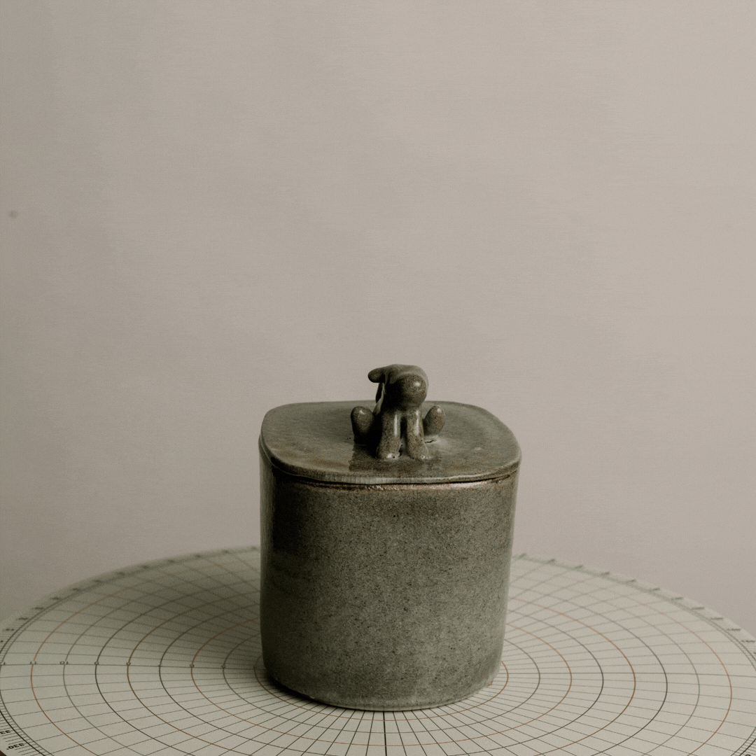 A ceramic container with a ceramic dog figurine on top, placed on a round grid-patterned surface against a plain background.