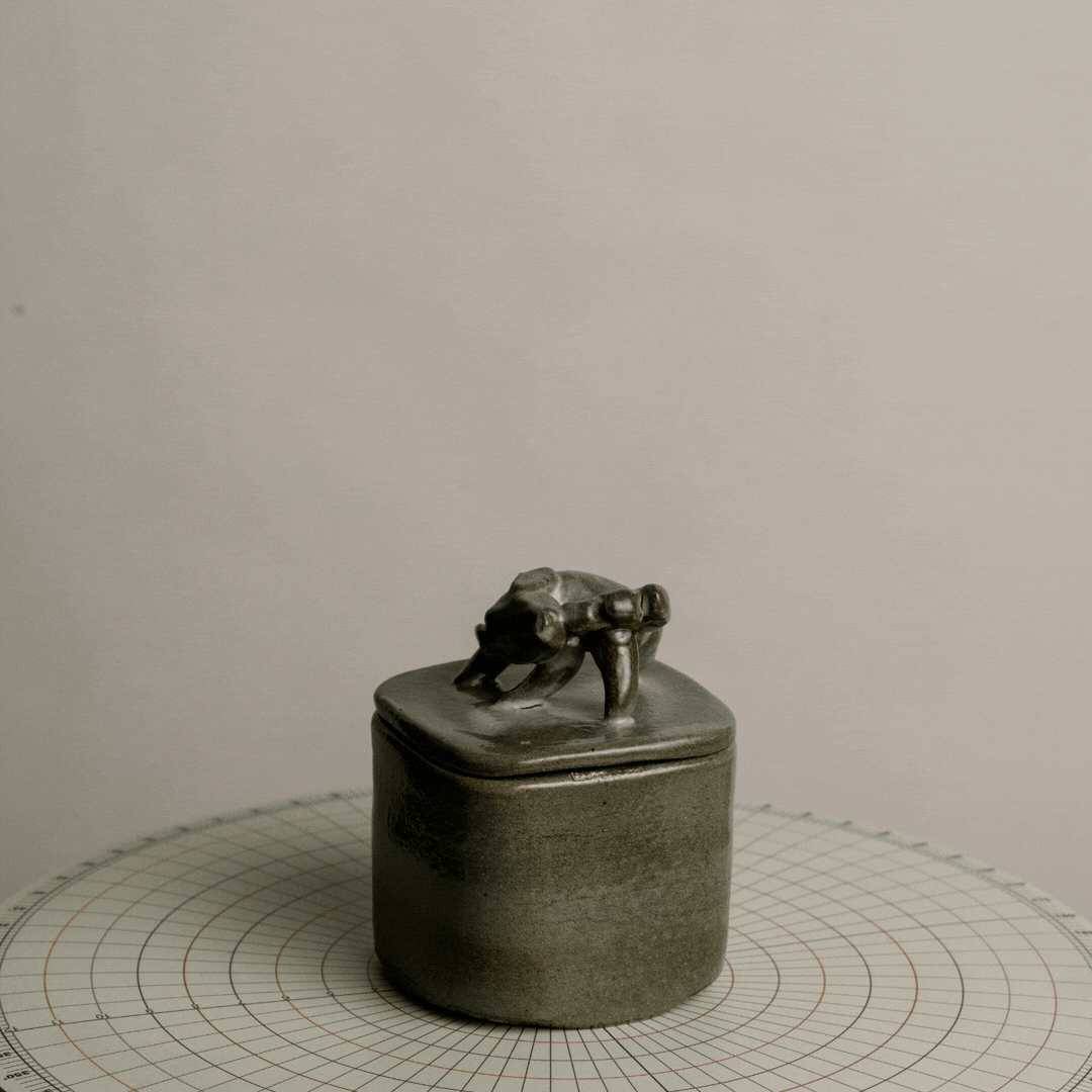 Small, dark ceramic container with a lid, topped with a sculpted figure resembling a frog or toad, sitting on a round, grid-lined surface with a plain, light-colored background.