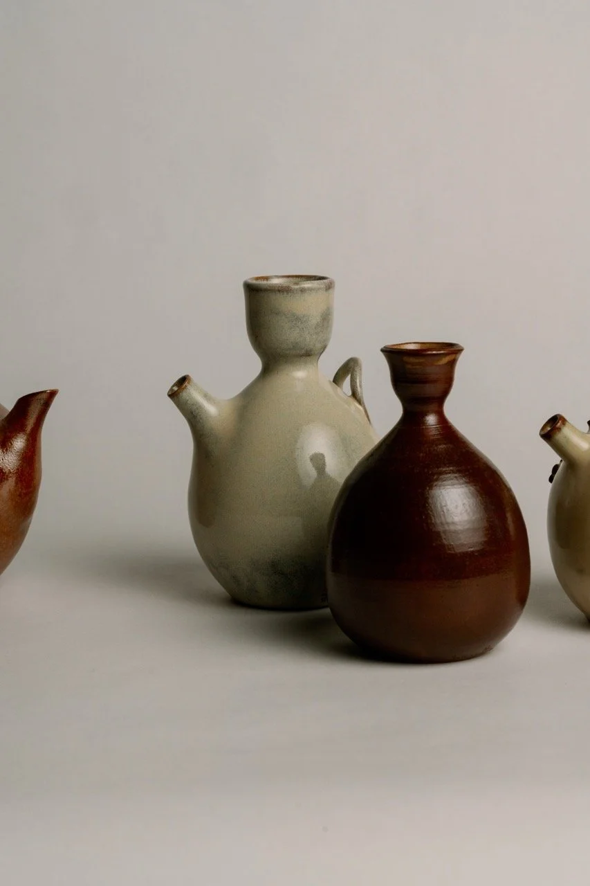 Several ceramic vases, including a greenish one with a lid and spout, a reddish-brown one with a narrow neck, and other partially visible vases, arranged on a plain surface against a neutral background.