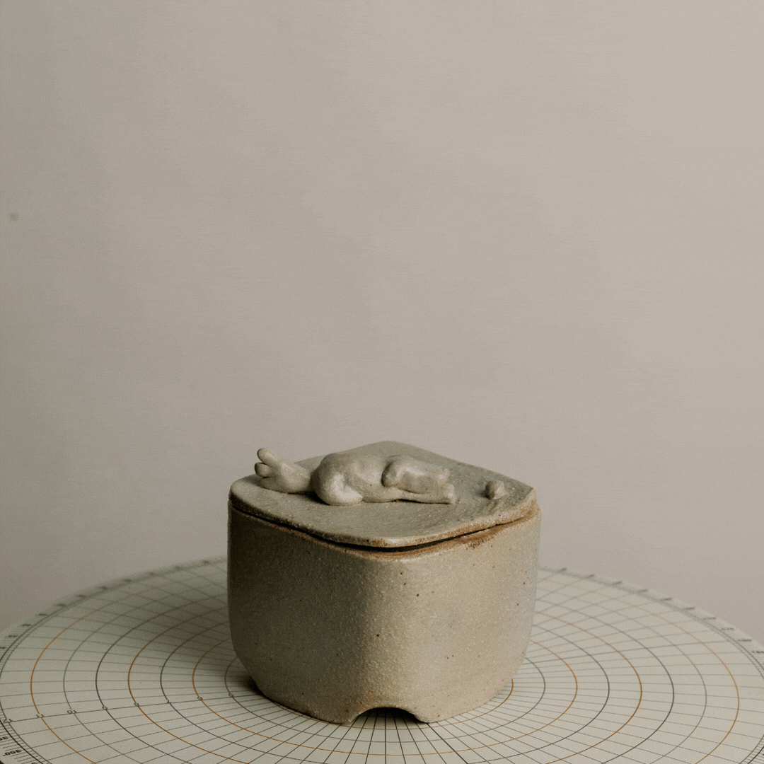 Small ceramic container with a lid, featuring a carved animal figure, placed on a measuring mat with grid lines, against a plain background.