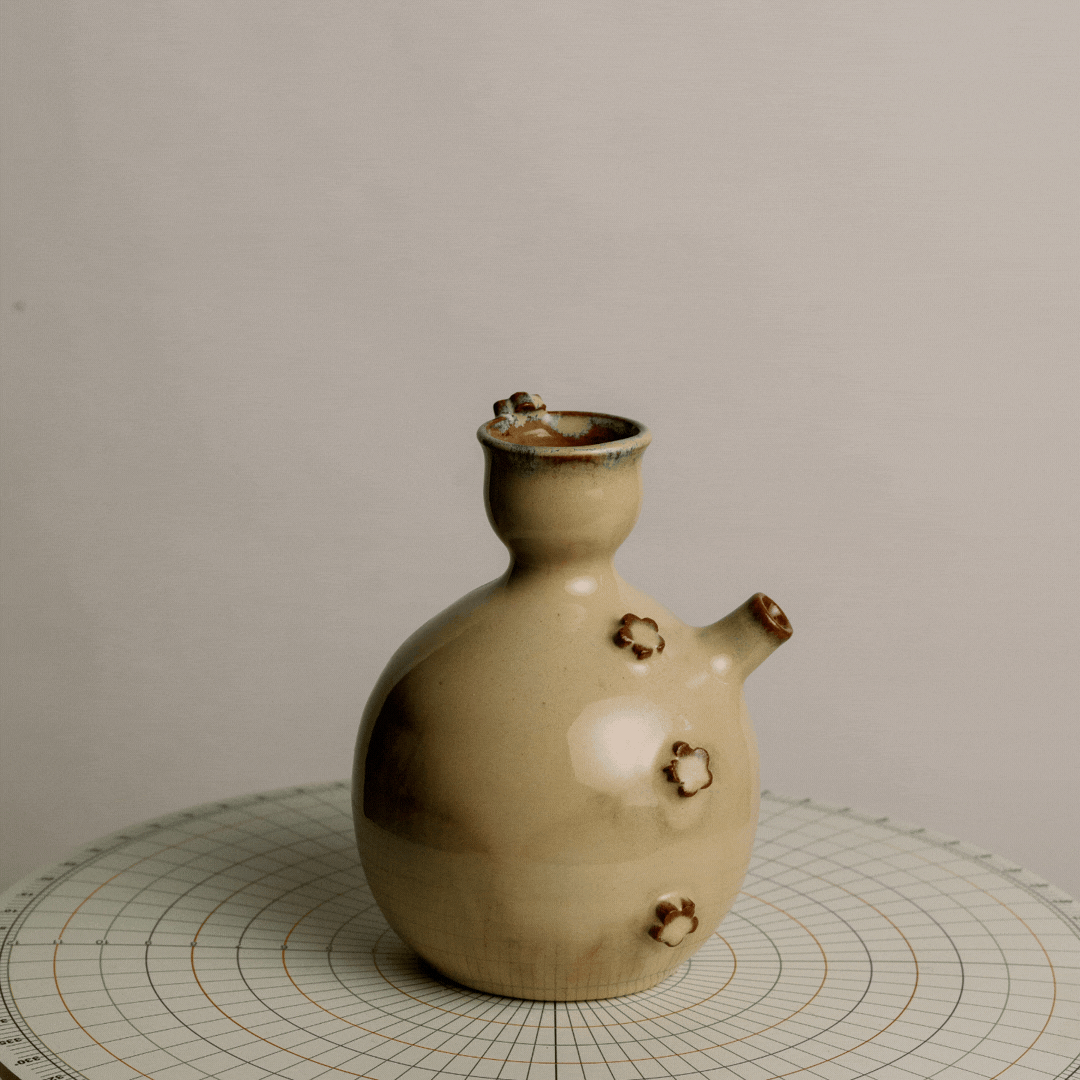 A beige ceramic vessel with a narrow neck and small flower-shaped details on its round body, placed on a round grid-patterned surface.