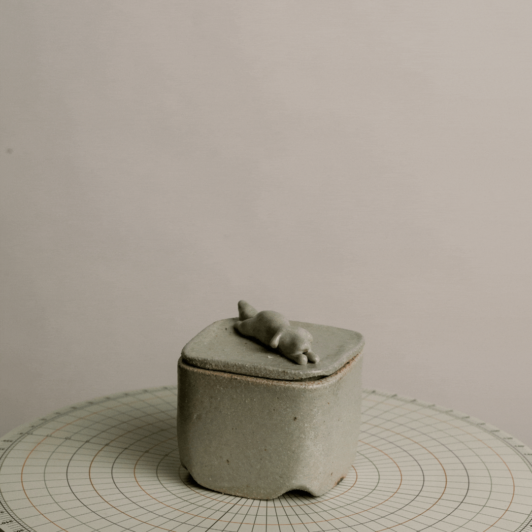 A small, rounded ceramic container with a lid, featuring a tiny animal figure on top, placed on a circular grid-patterned surface against a plain background.