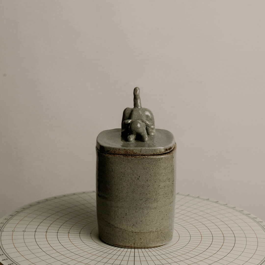 Gray ceramic container with a lid, topped by a small sculpture of an elephant with a long trunk, placed on a circular measuring mat.