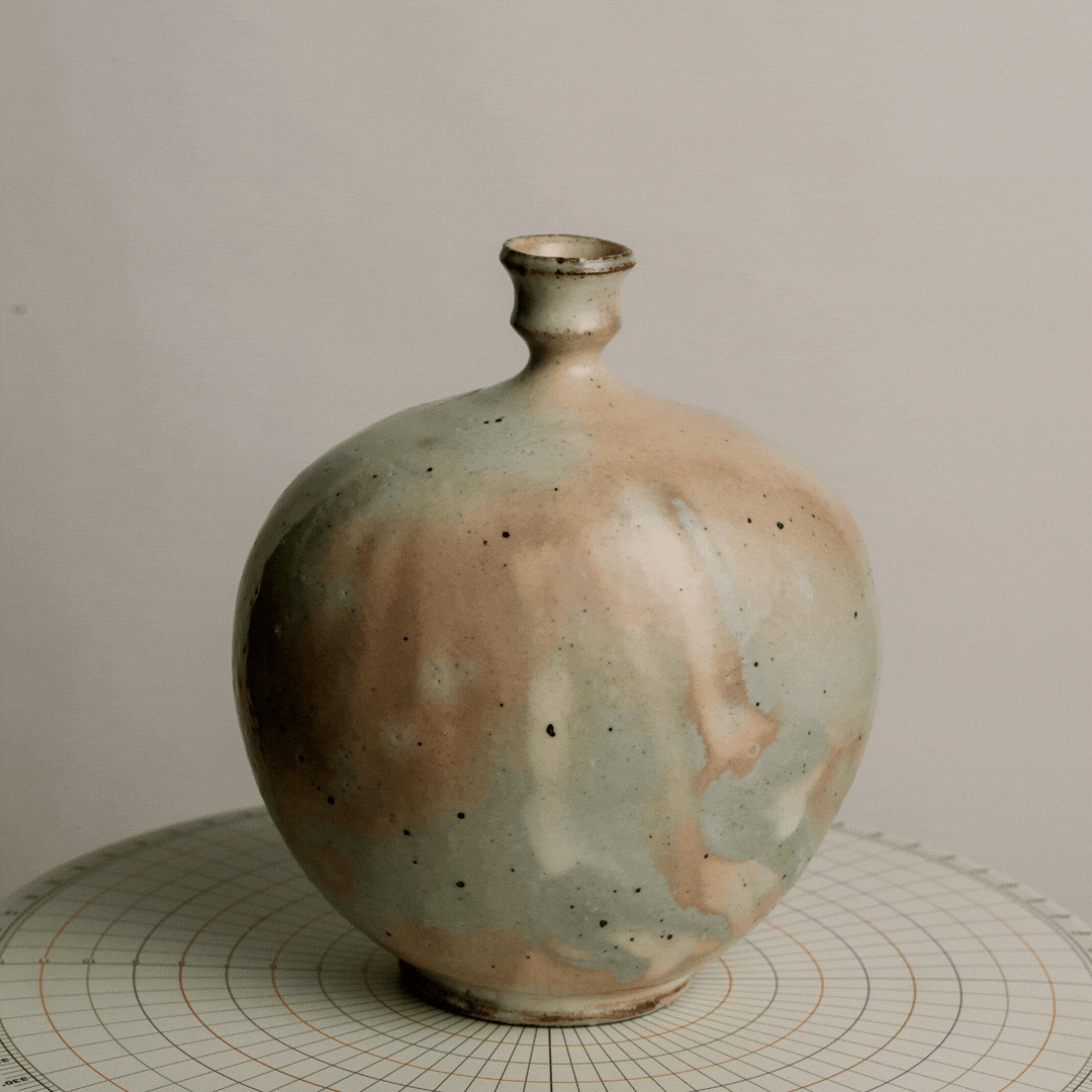 A small ceramic vase with a narrow neck and rounded body, glazed in soft pastel colors with a speckled pattern, displayed on a circular grid-patterned surface.