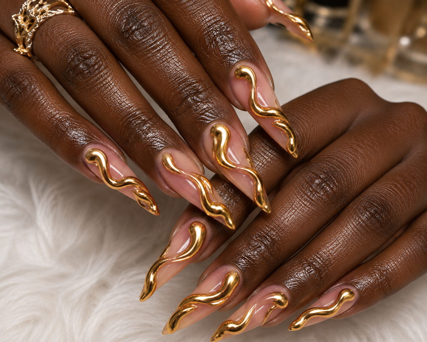 Close-up of two hands with dark skin, showing long, wavy, gold-colored nail art and gold rings on fingers, resting on a soft white surface.