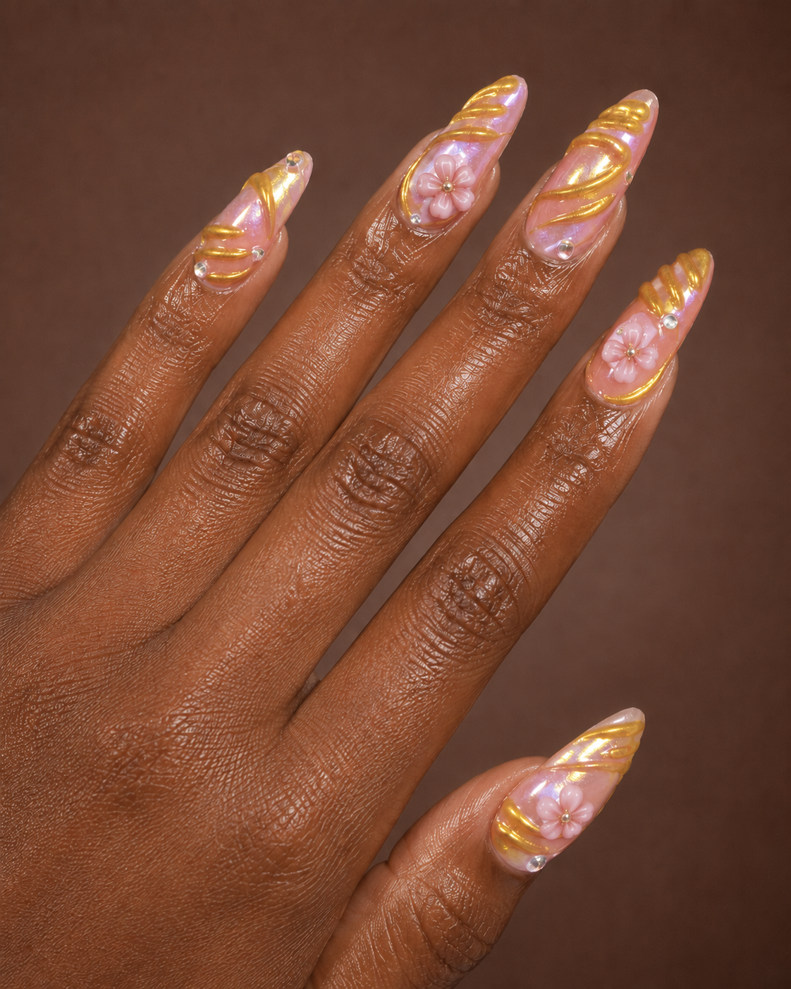 Close-up of a hand with decorated long acrylic nails featuring pink and gold swirl designs, small pink flowers with tiny pearls, and clear rhinestones on a brown background.