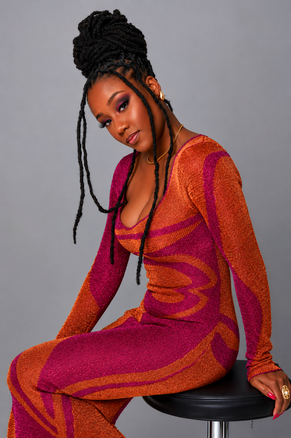 A woman with dark skin and long twisted hair styled in an updo, wearing colorful makeup, gold jewelry, and a vibrant, patterned, long-sleeved dress in shades of orange and pink, sitting on a stool against a gray background.