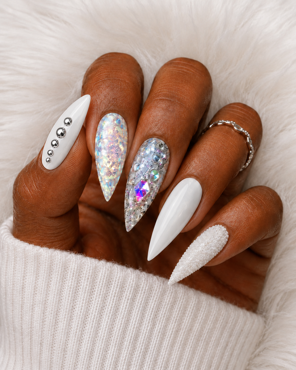 Close-up of a hand with long decorated nails featuring rhinestones, holographic designs, and white polish, resting on light-colored fabric.