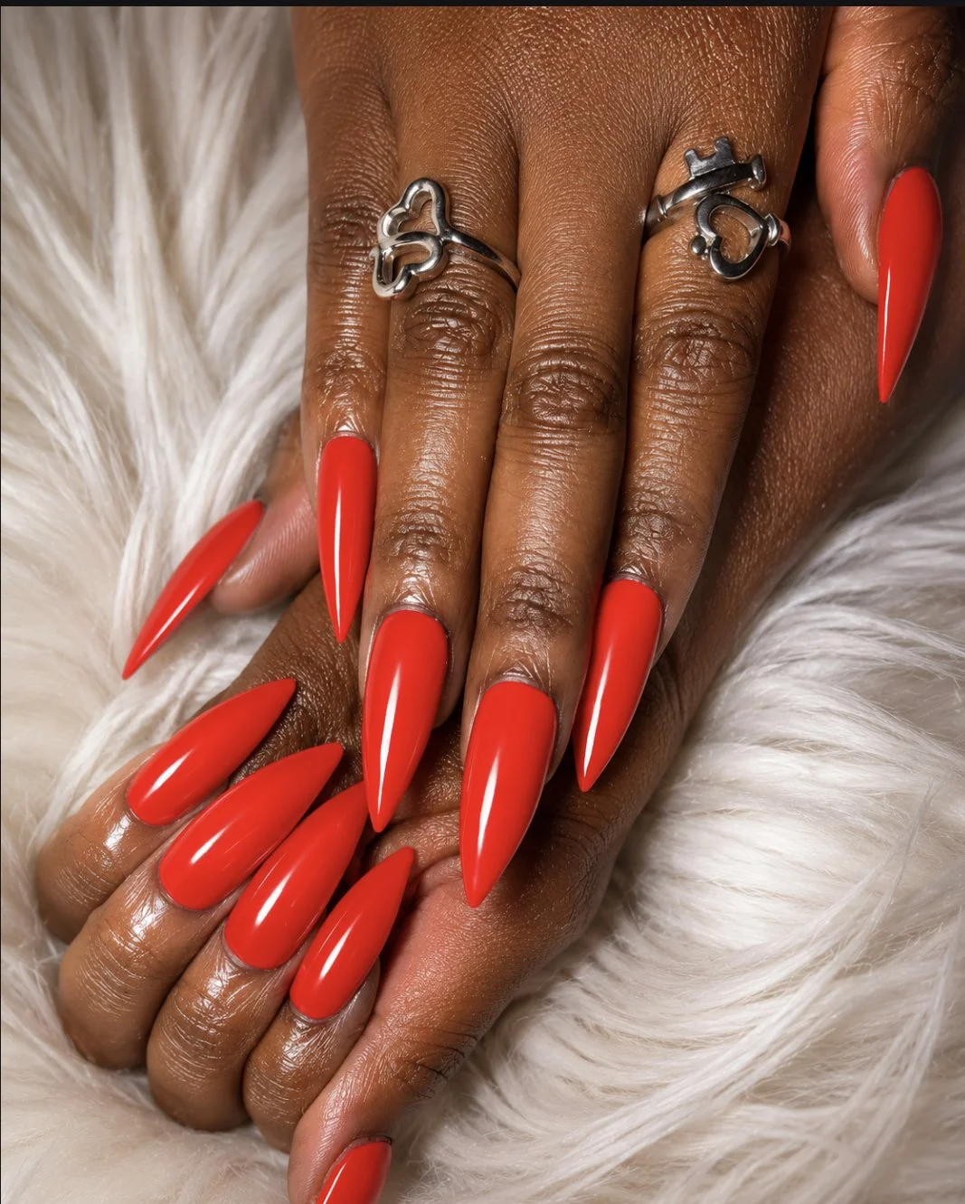 Close-up of a hand with long, pointed red nails, resting on a white furry surface, wearing a silver ring with a heart and lock design.