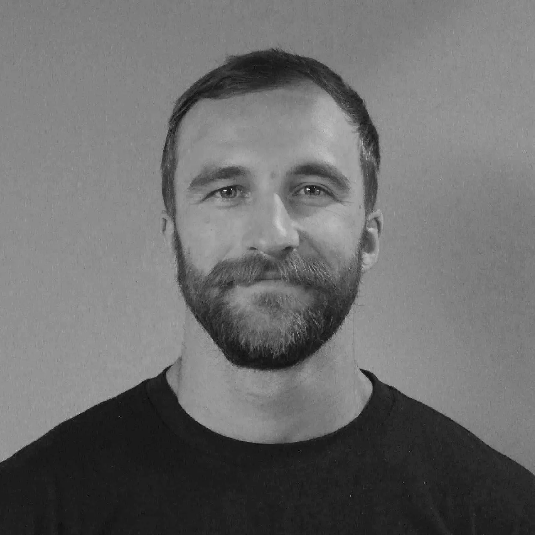 Black and white portrait of a man with short dark hair, a beard, and wearing a black shirt, smiling slightly against a plain wall background.