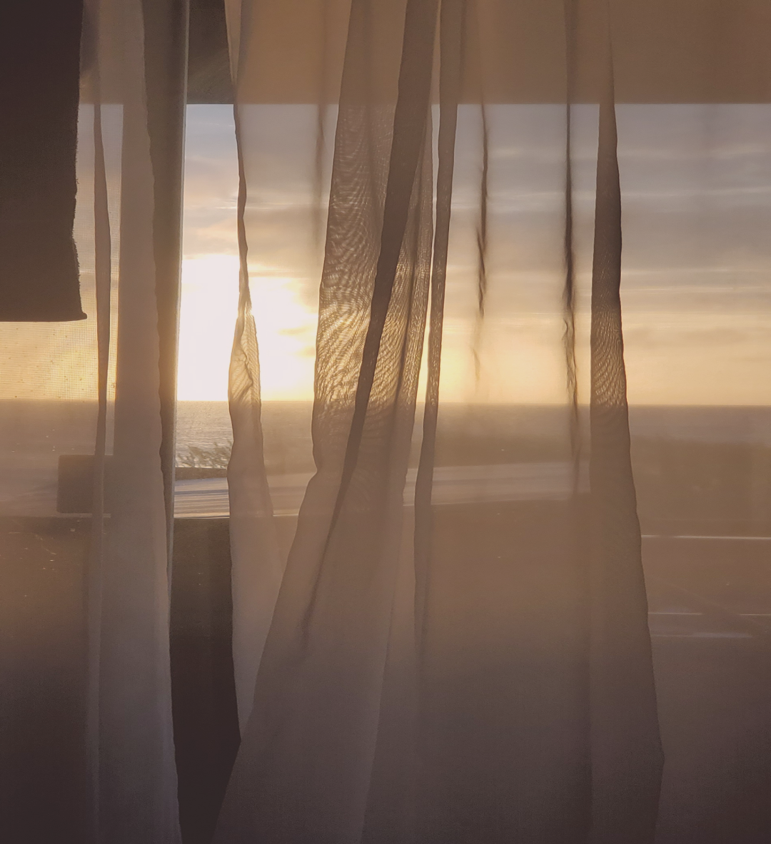 Sunset view through sheer curtains overlooking the ocean.
