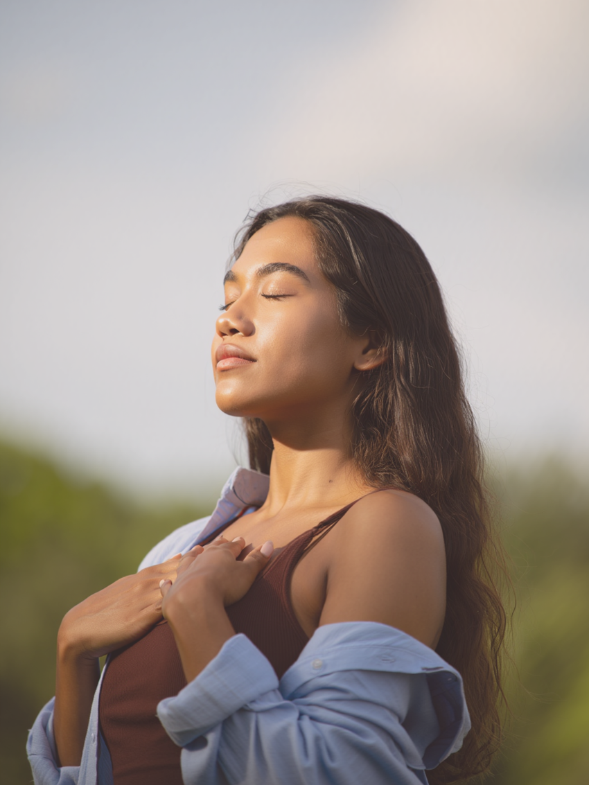 A woman with long hair standing outdoors with eyes closed, holding her coat open over her chest, enjoying sunlight.