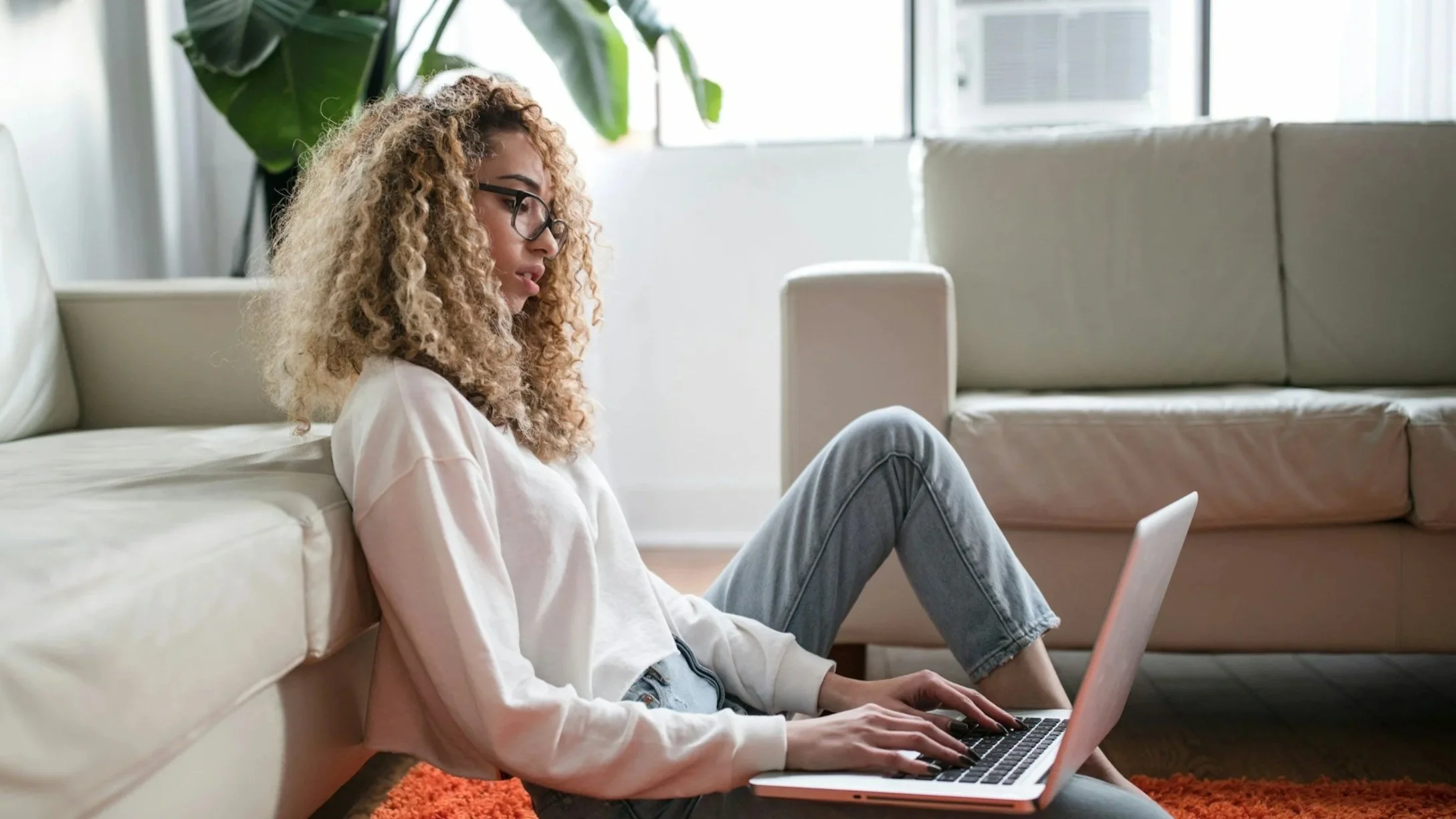 A young woman with curly blonde hair and glasses sits on the floor beside a white sofa, using a laptop in a bright, modern living room with large windows and a green plant in the background.