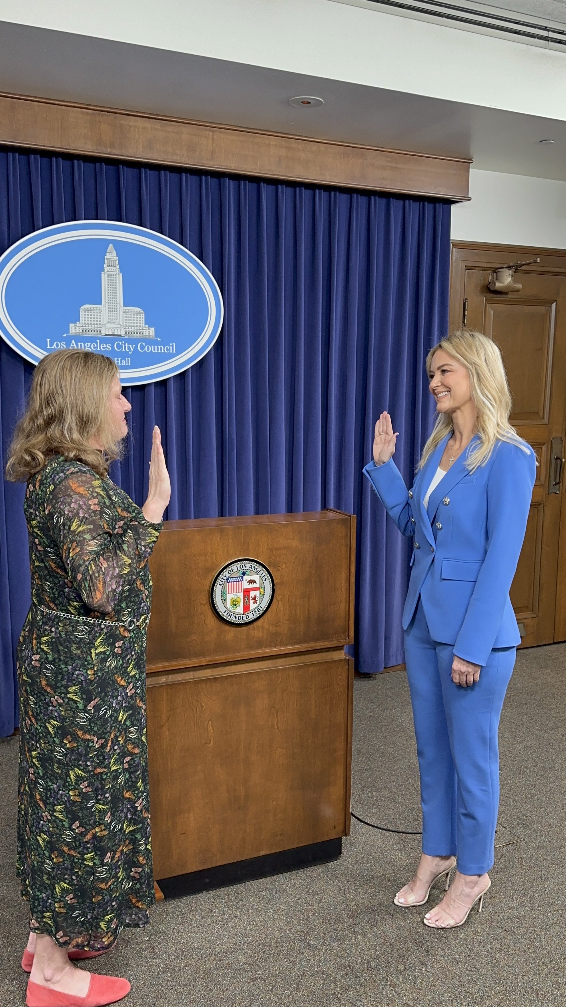 Two women standing for a swearing-in ceremony at Los Angeles City Hall, raising their right hands. The woman on the right is dressed in a bright blue pantsuit and beige high heels, smiling. The woman on the left is wearing a floral dress with pink shoes. A blue curtain, the Los Angeles City Council sign, and the city emblem are visible behind them.