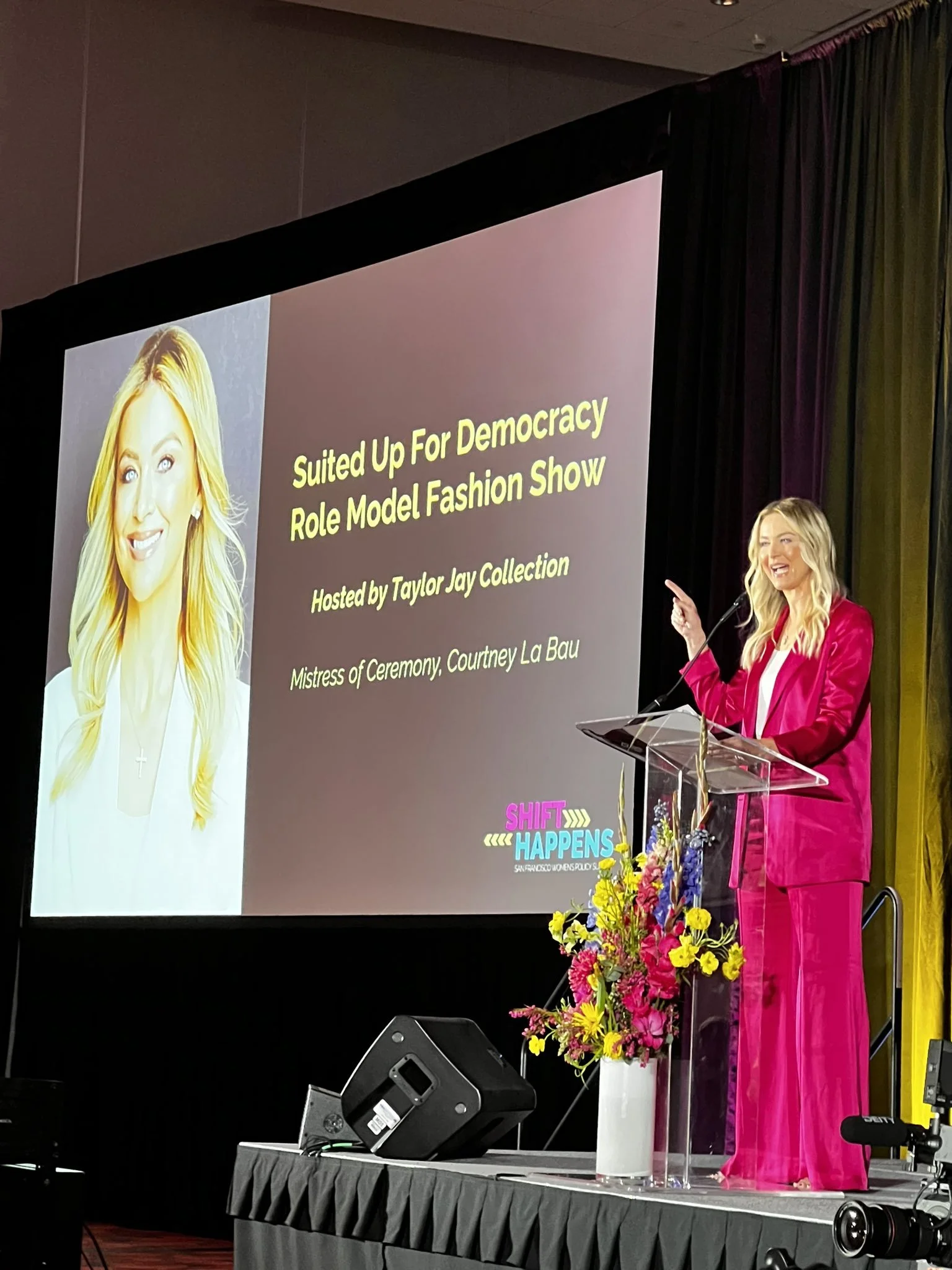 Courtney La Bau in a bright pink suit is speaking at a podium with a large screen behind her displaying a presentation slide. The slide shows a portrait of a woman with long blonde hair,  and hosting and MC'ing an event. Courtney La Bau.