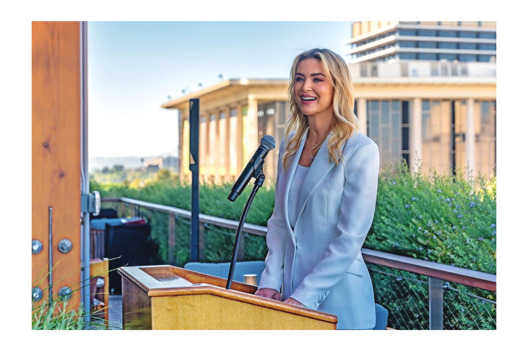 A woman with blonde hair, dressed in a white blazer, stands at a podium outdoors, smiling, speaking into a microphone. In the background, there are modern buildings and lush greenery. Courtney La Bau.  JPMorgan.