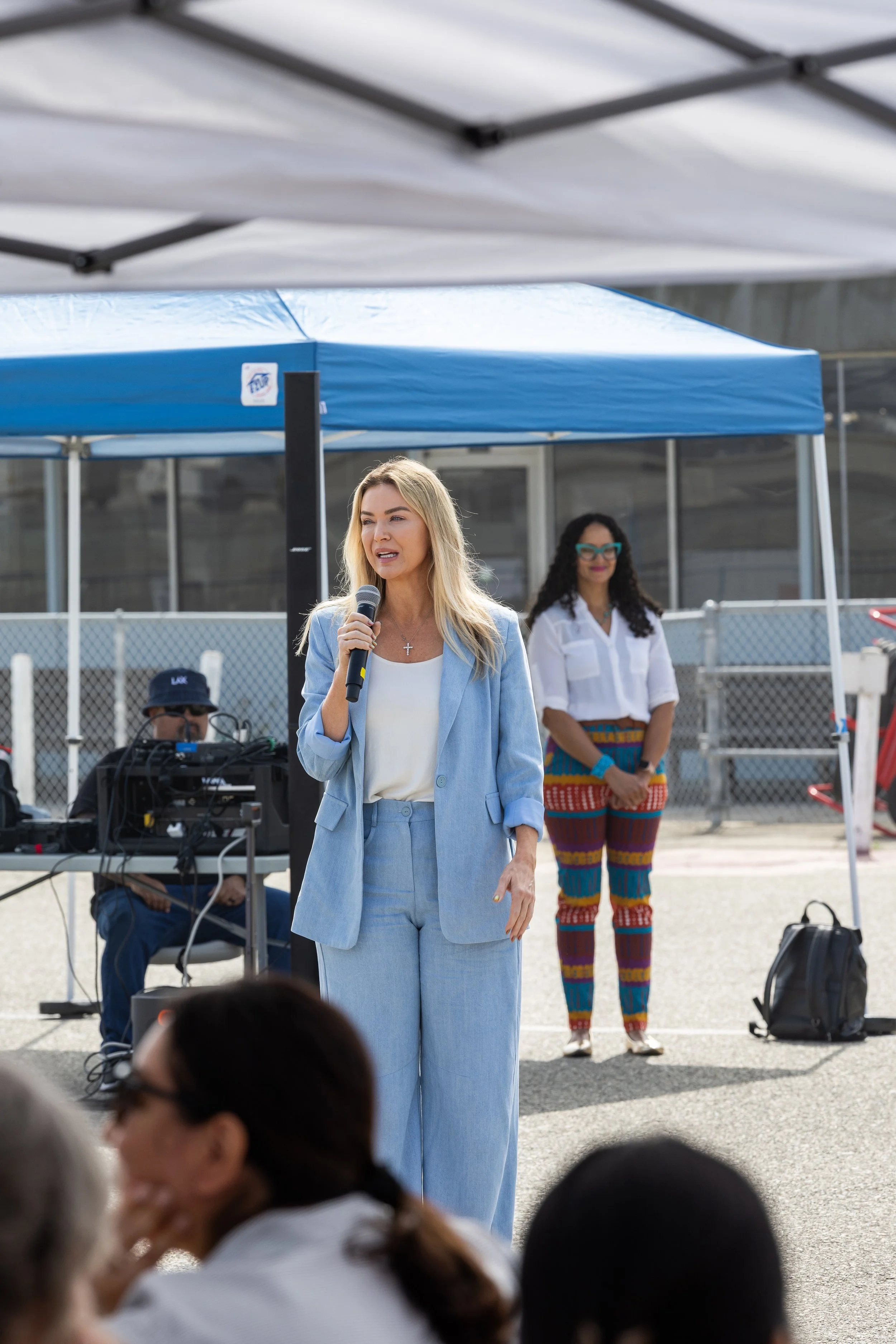 A woman with long blonde hair in a blue suit speaking into a microphone at an outdoor event, with people and tents in the background for Los Angeles World Airports, LAX, women in leadership, Commissioner, Executive.