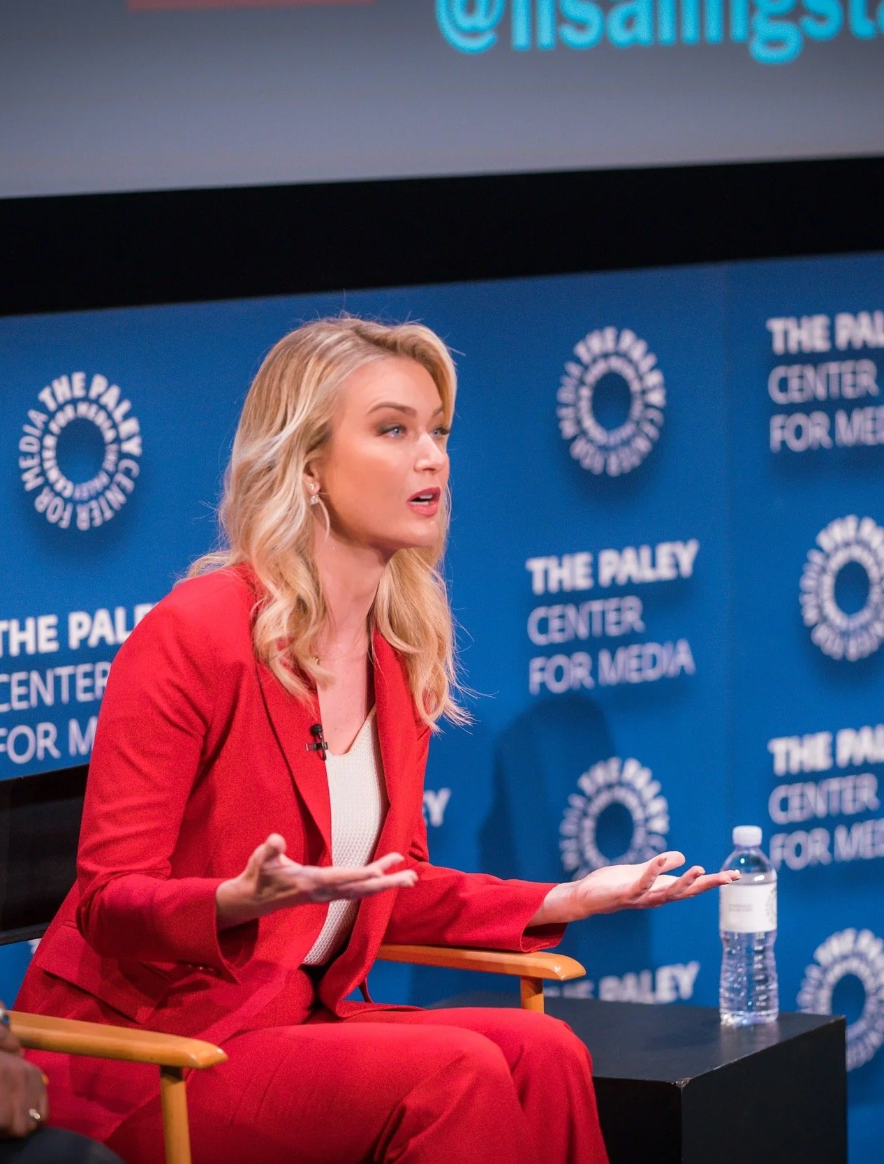 A woman with blonde hair in a red blazer and red pants is speaking during a panel at The Paley Center for Media. She has her hands open and is gesturing as she speaks, smart and classy and a leader in media and speaking pundit.