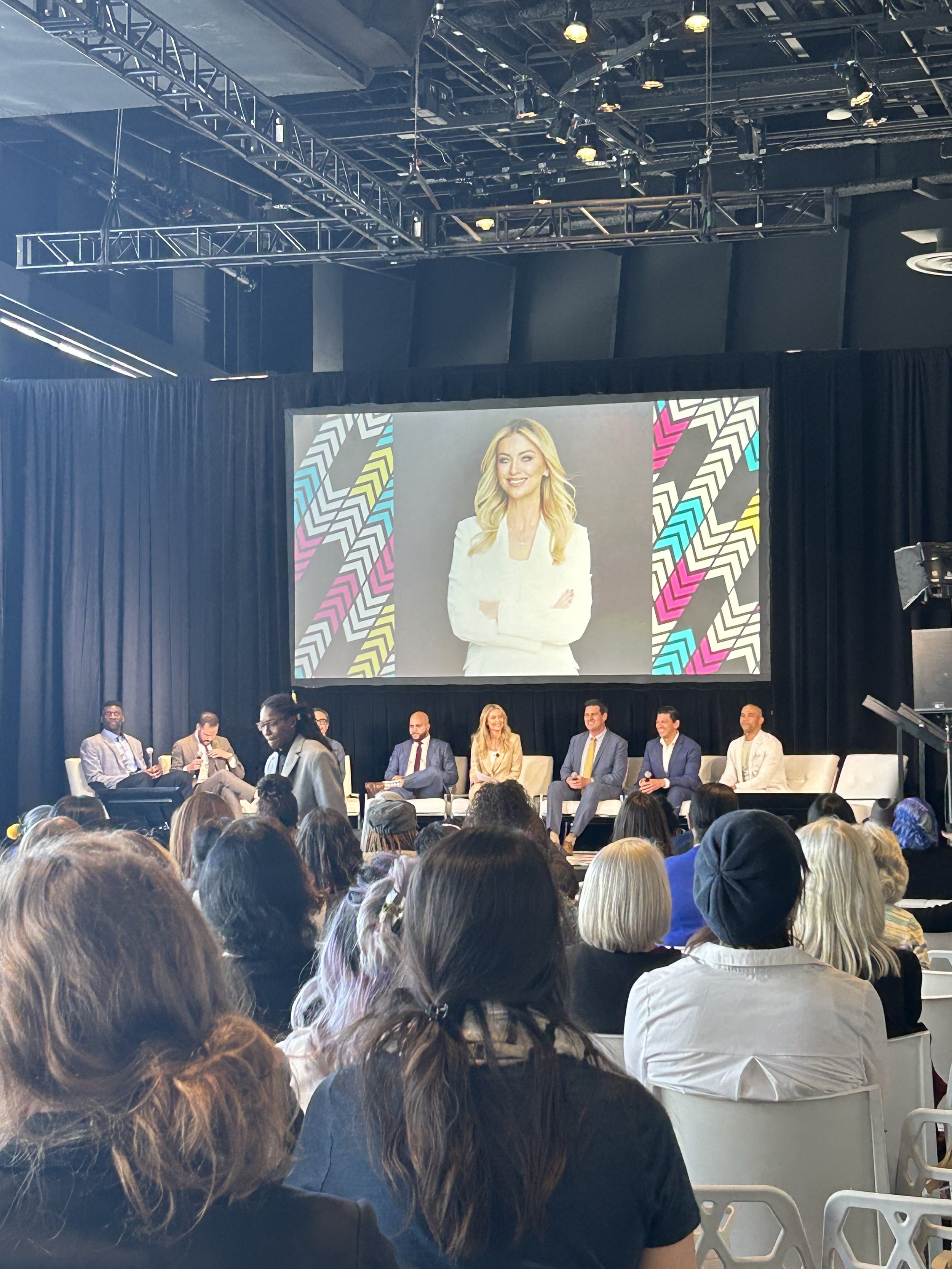 Panel of seven people seated on stage during conference or panel discussion, audience in foreground, large screen behind displaying a woman in white blazer with crossed arms, colorful geometric background.