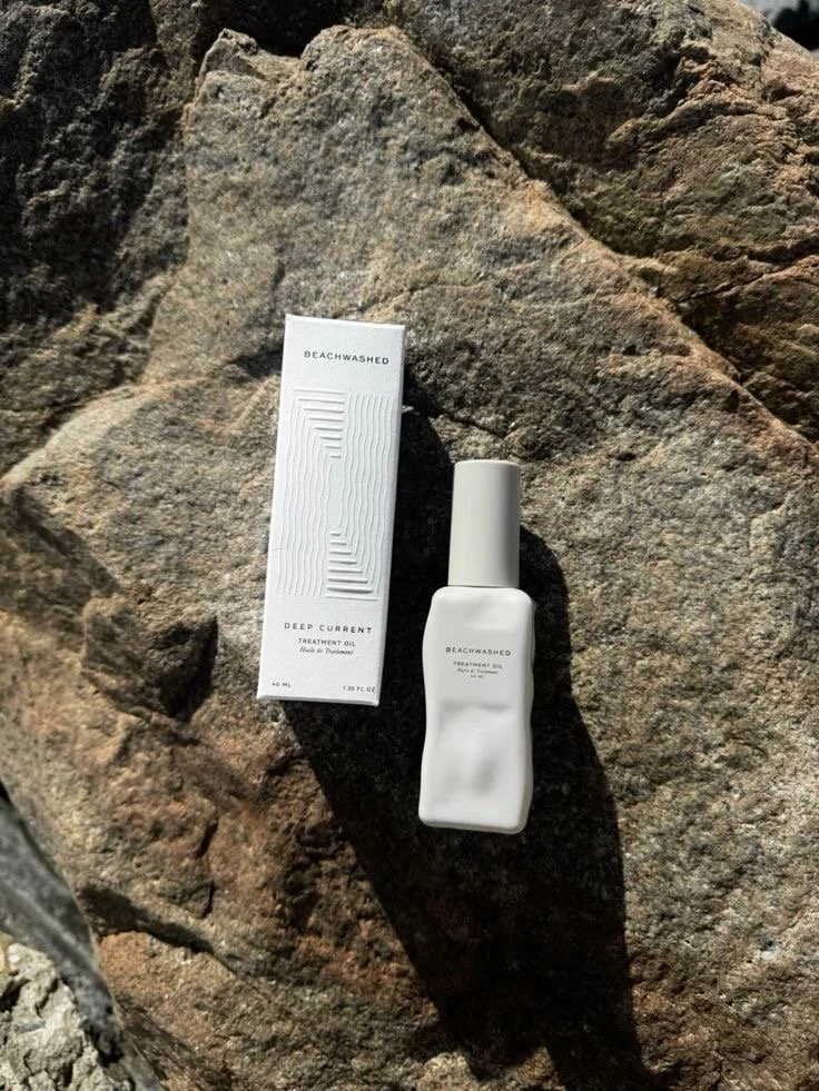 A white bottle and a white box placed on a large rock outdoors. The bottle is labeled 'BEACHWASHED Treatment Oil' and the box has 'BEACHWASHED Deep Current Treatment Oil' printed on it.