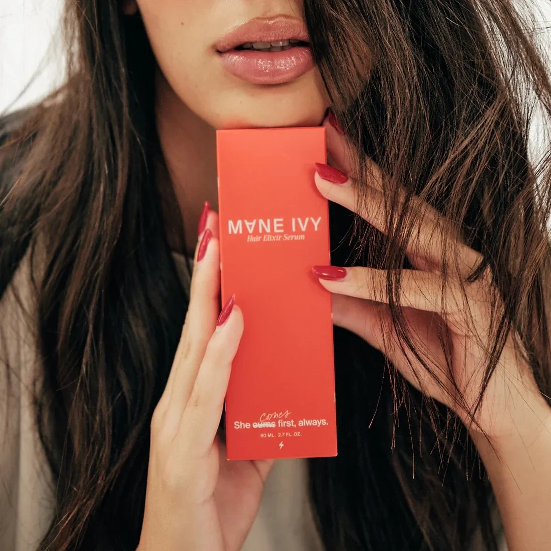 A woman with long brown hair and red nail polish holding a red box of Mane Ivy Hair Elixir Serum near her face, with her lips slightly parted.
