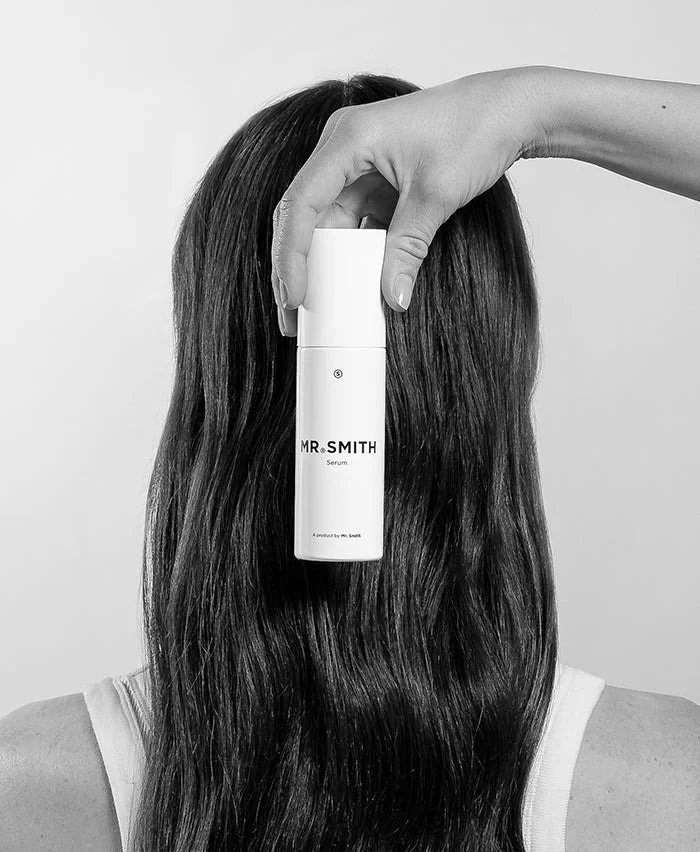 A person with long, dark hair holds a serum bottle labeled 'MR. SMITH' in front of their face, obscuring it. The background is plain and the image is in black and white.