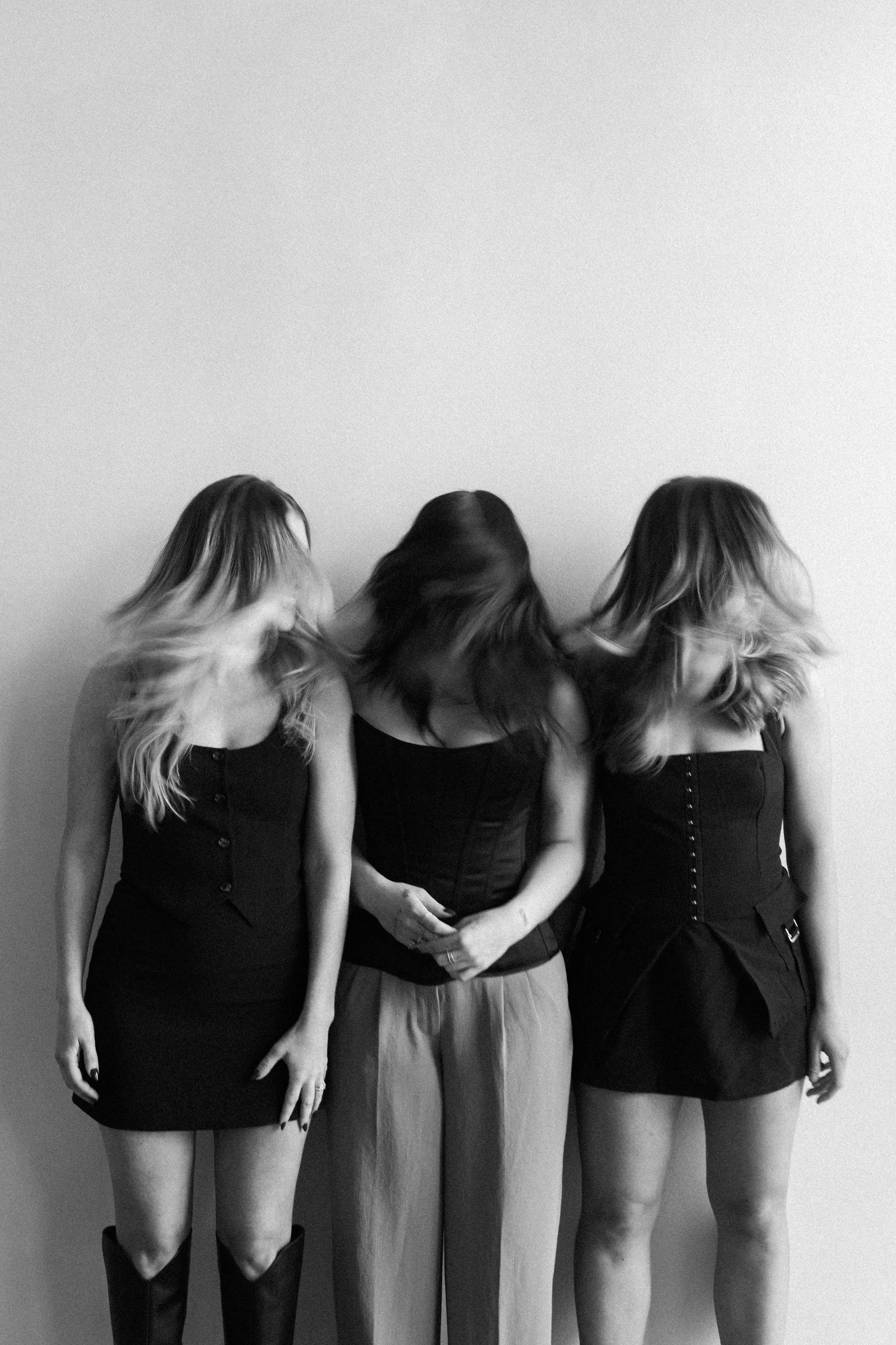 Three women standing side by side, with their heads turned to the left, obscuring their faces. They are dressed in black and gray, with long hair, in front of a plain wall.