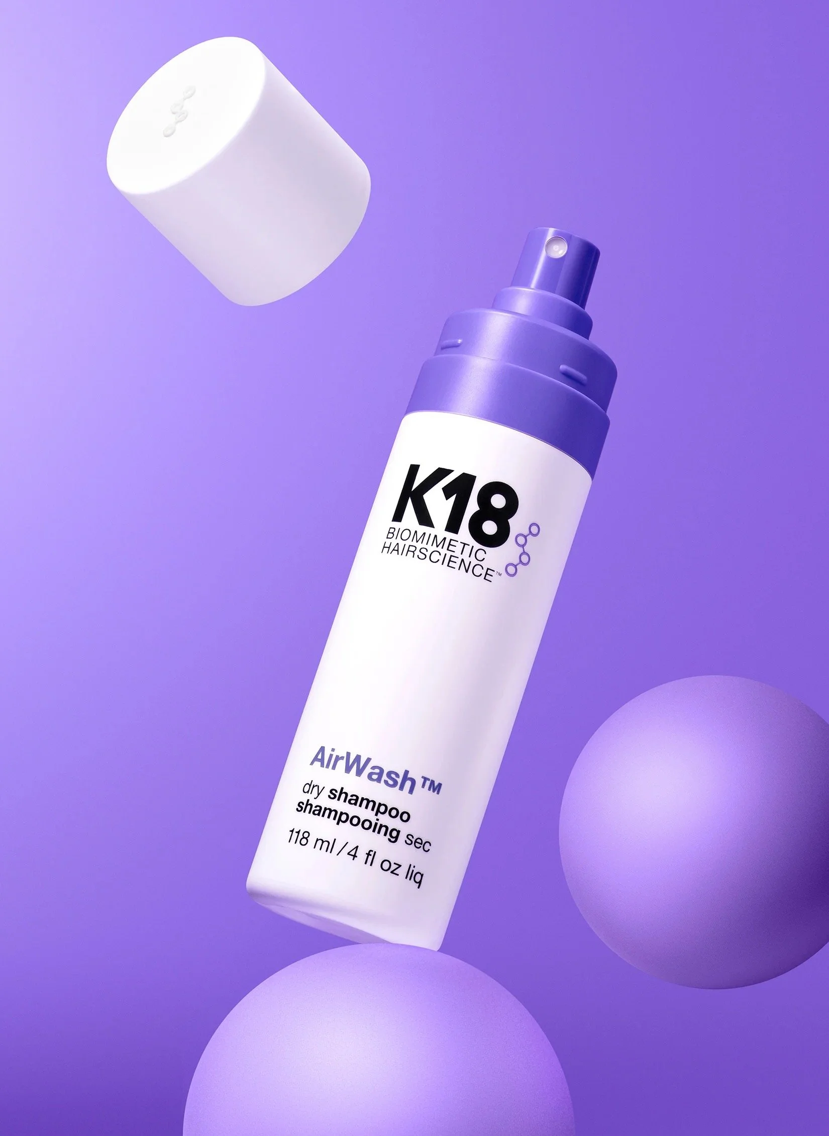 A white bottle of K18 Biomimetic Hairscience AirWash dry shampoo with a purple cap is shown against a purple background, with the cap placed near the top left of the bottle.