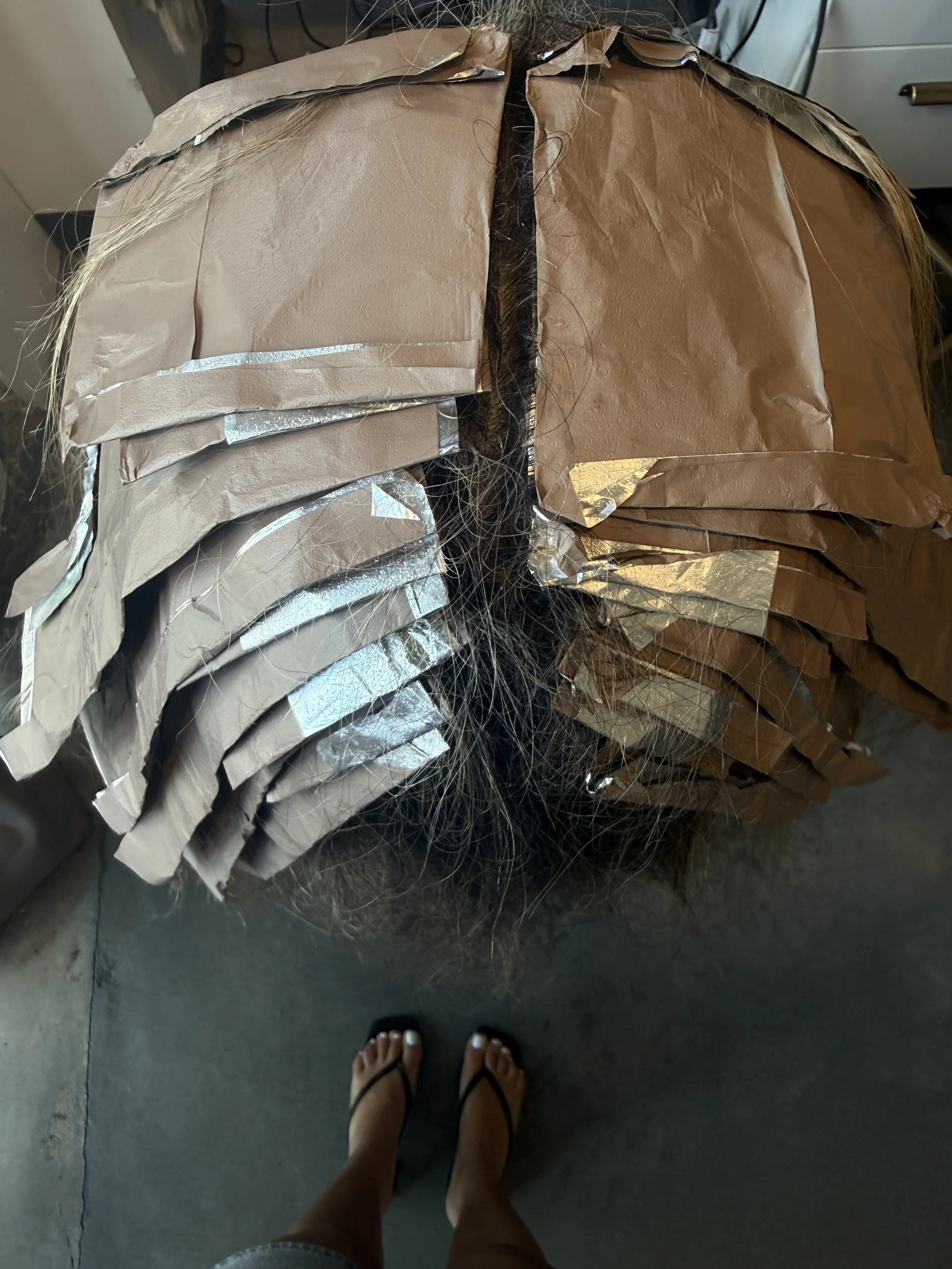 Top view of a person standing on a concrete floor, wearing flip-flops, looking at a large pile of human hair wrapped in brown paper with silver tape, possibly hair extensions or wigs.