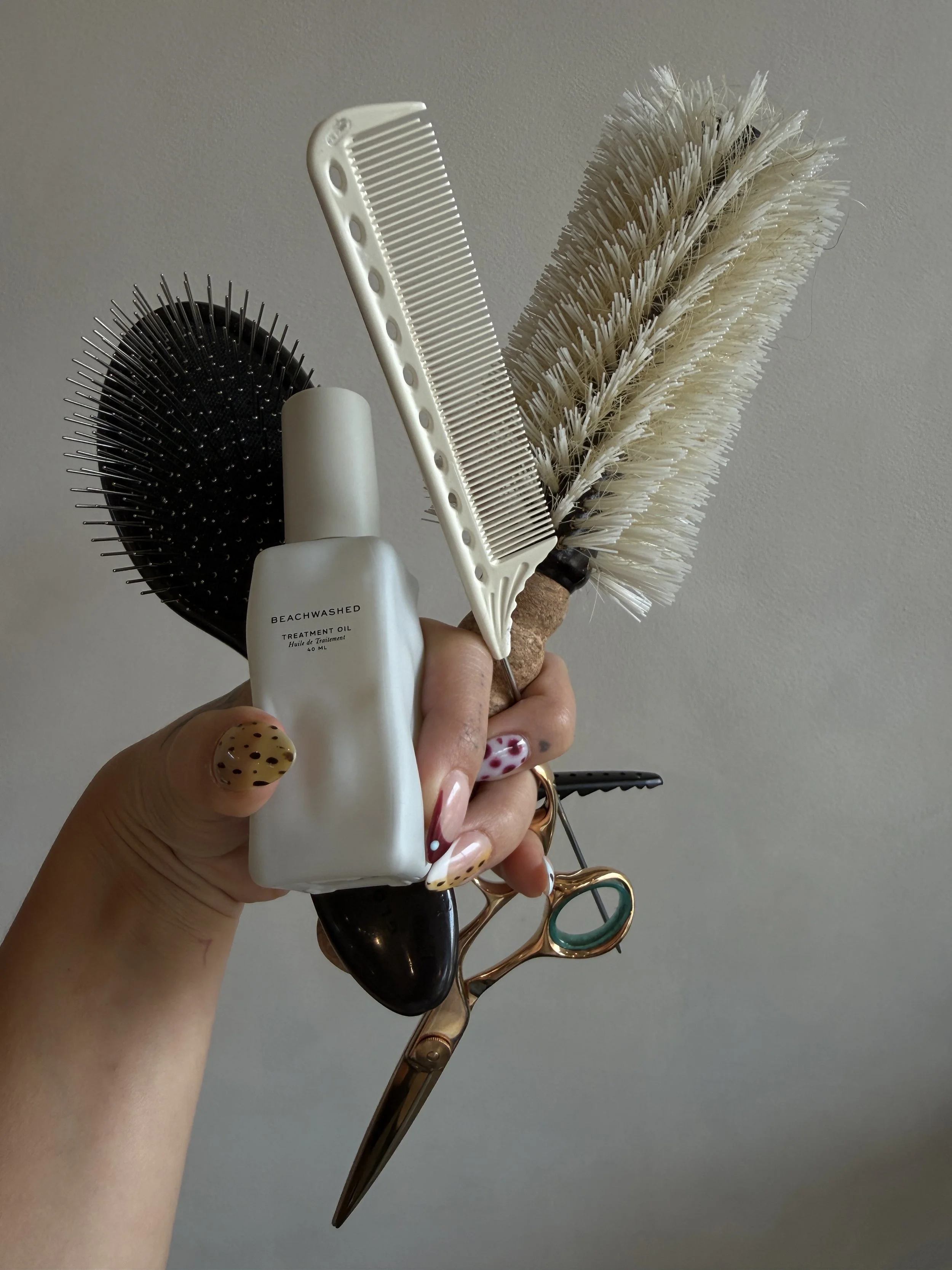 A hand holding a collection of hair styling tools, including a round brush, a wide-tooth comb, a small spike brush, and a pair of hair scissors.