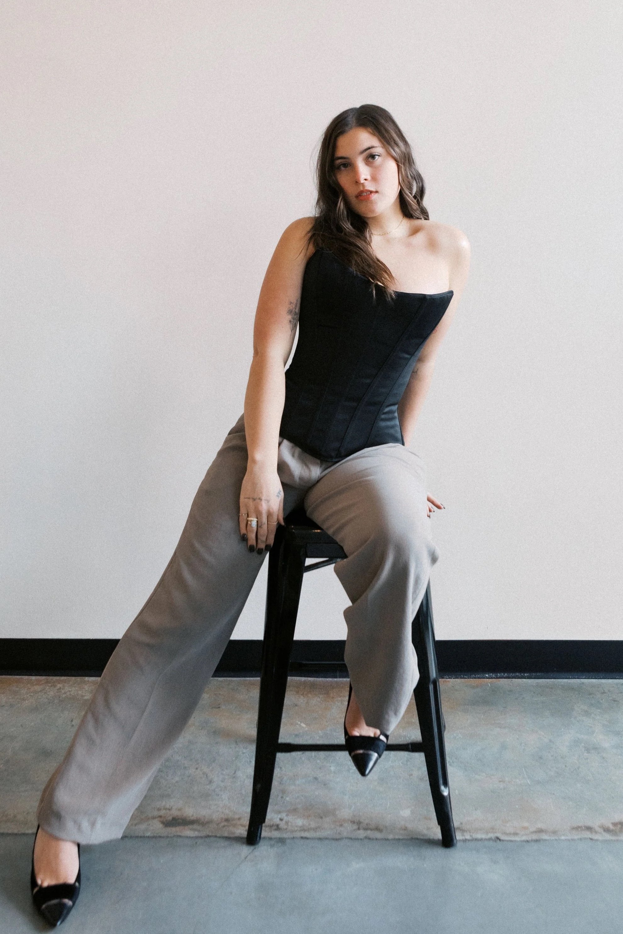 A young woman with brown wavy hair sitting on a black stool against a plain light-colored wall. She is wearing a black strapless corset top, beige wide-leg pants, and black pointed-toe heels. She has rings on her fingers and tattoos on her arm and hand.