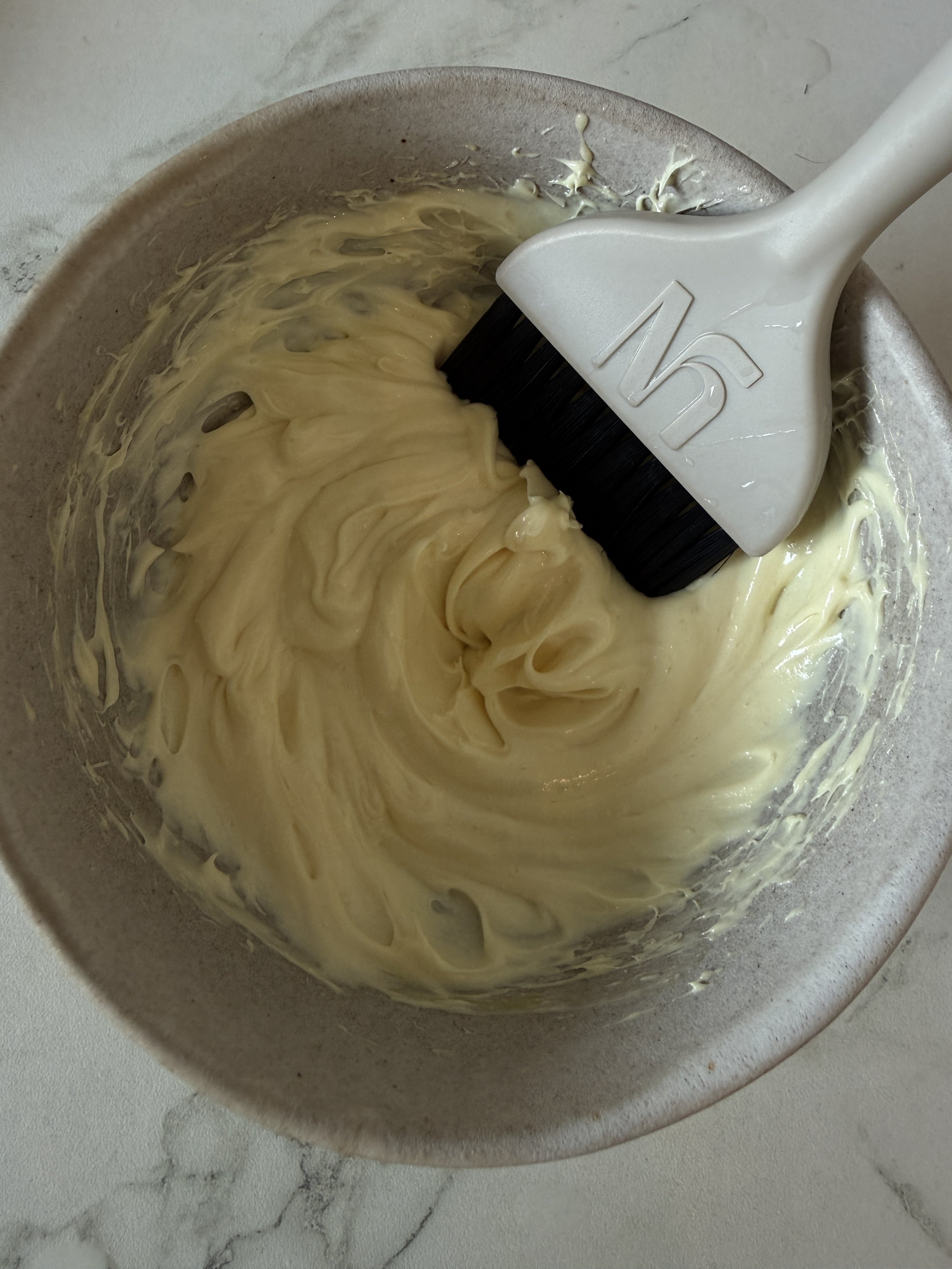 A bowl with a white hand mixer beaters attached, containing a creamy, pale yellow mixture, sitting on a light-colored marble surface.