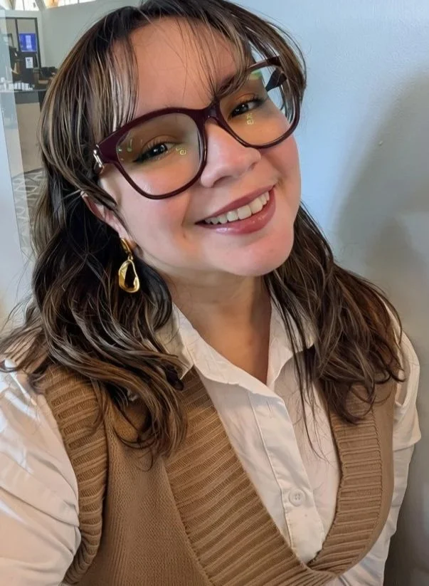 A young woman with wavy brown hair, wearing large glasses with a red frame, gold hoop earrings, a white collared shirt, and a beige vest, smiling indoors.