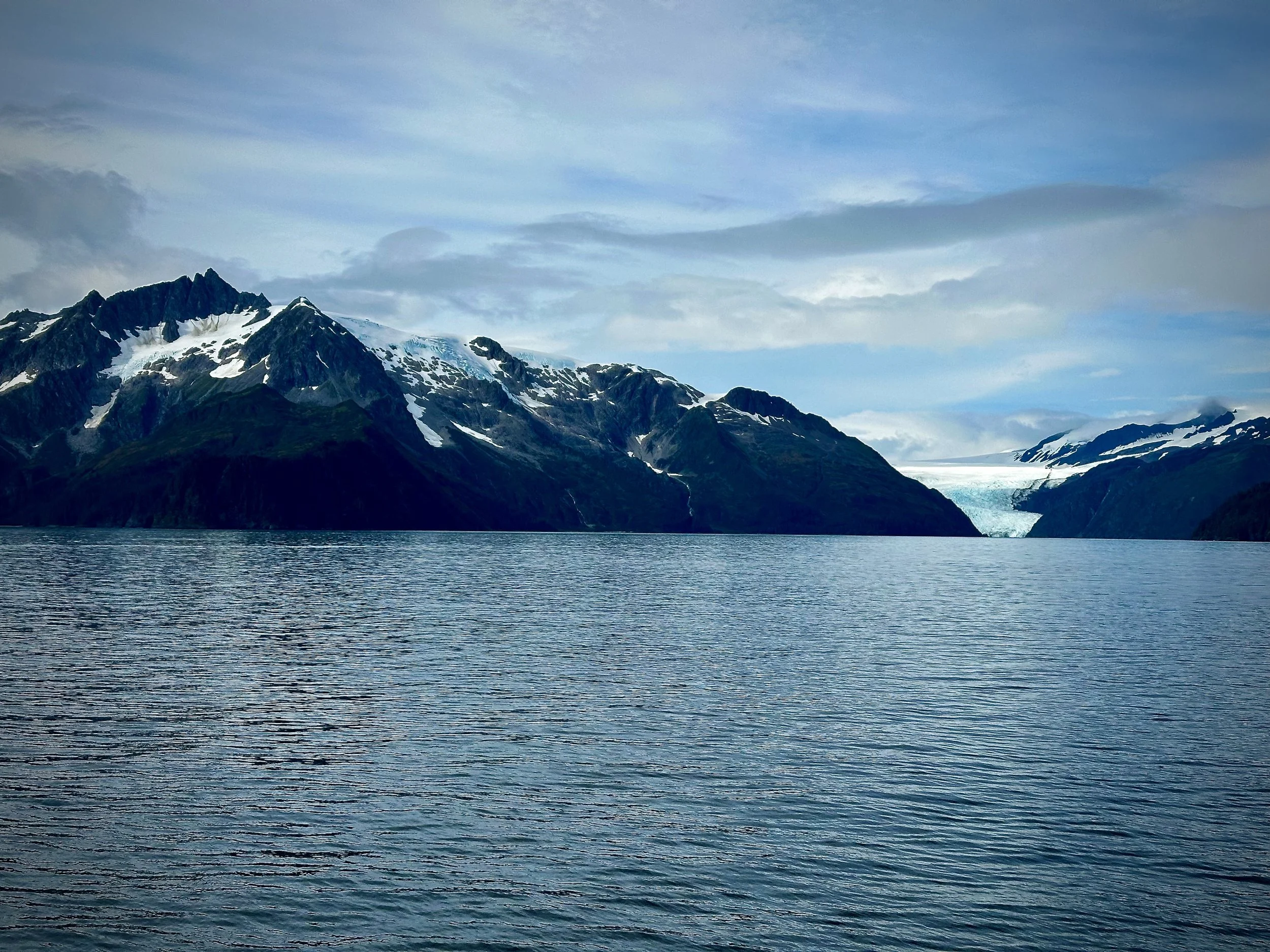 Snow-capped mountains with a glacier visible between peaks, viewed from the water in Kenai Fjords, Alaska