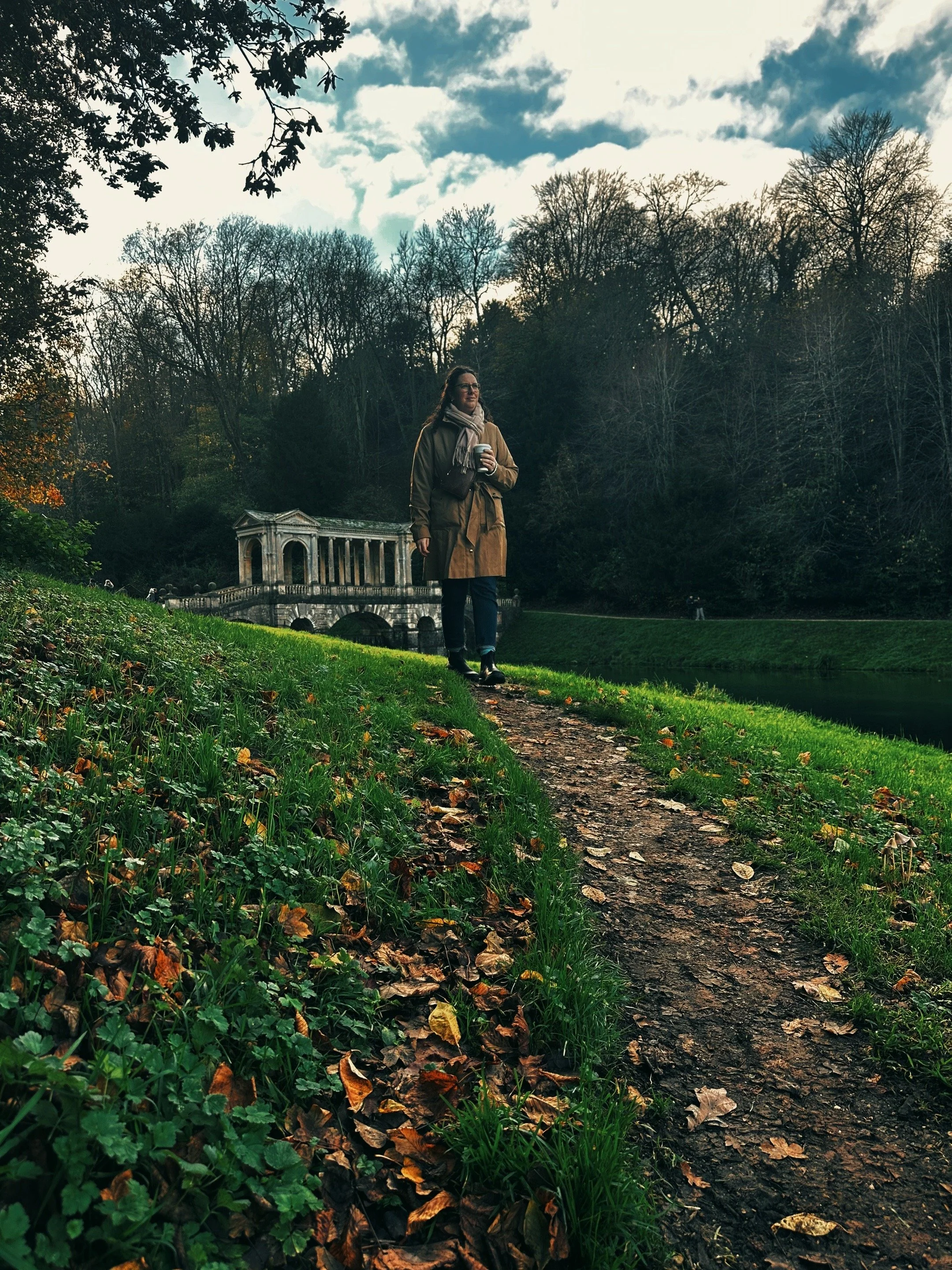 A woman walking on a dirt path beside a river in a park during fall, with trees and a stone bridge in the background, under a partly cloudy sky.