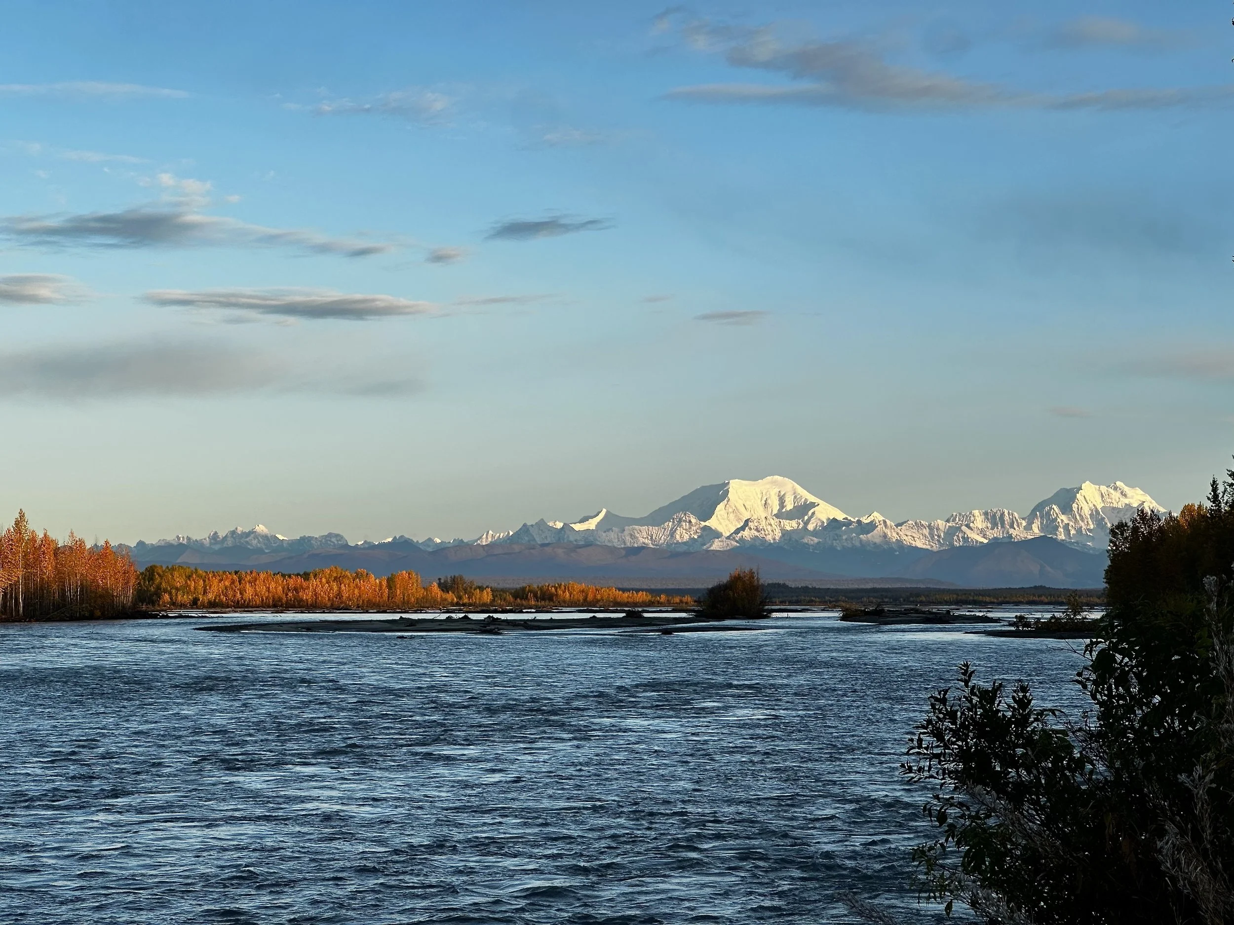 Scenic landscape featuring a river in the foreground, a line of trees with autumn foliage, snow-capped mountains in the distance, and a blue sky with scattered clouds.
