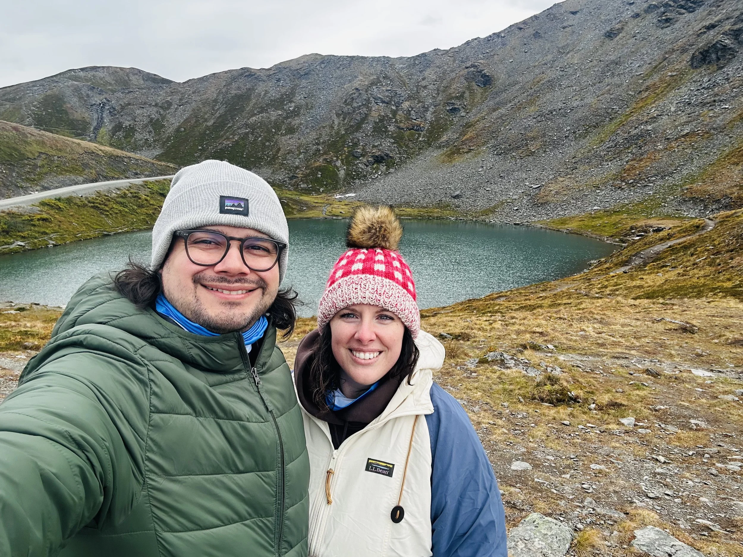 Jen and Shane smiling at Summit Lake along Hatcher's Pass, with rocky mountains and a winding road in the background