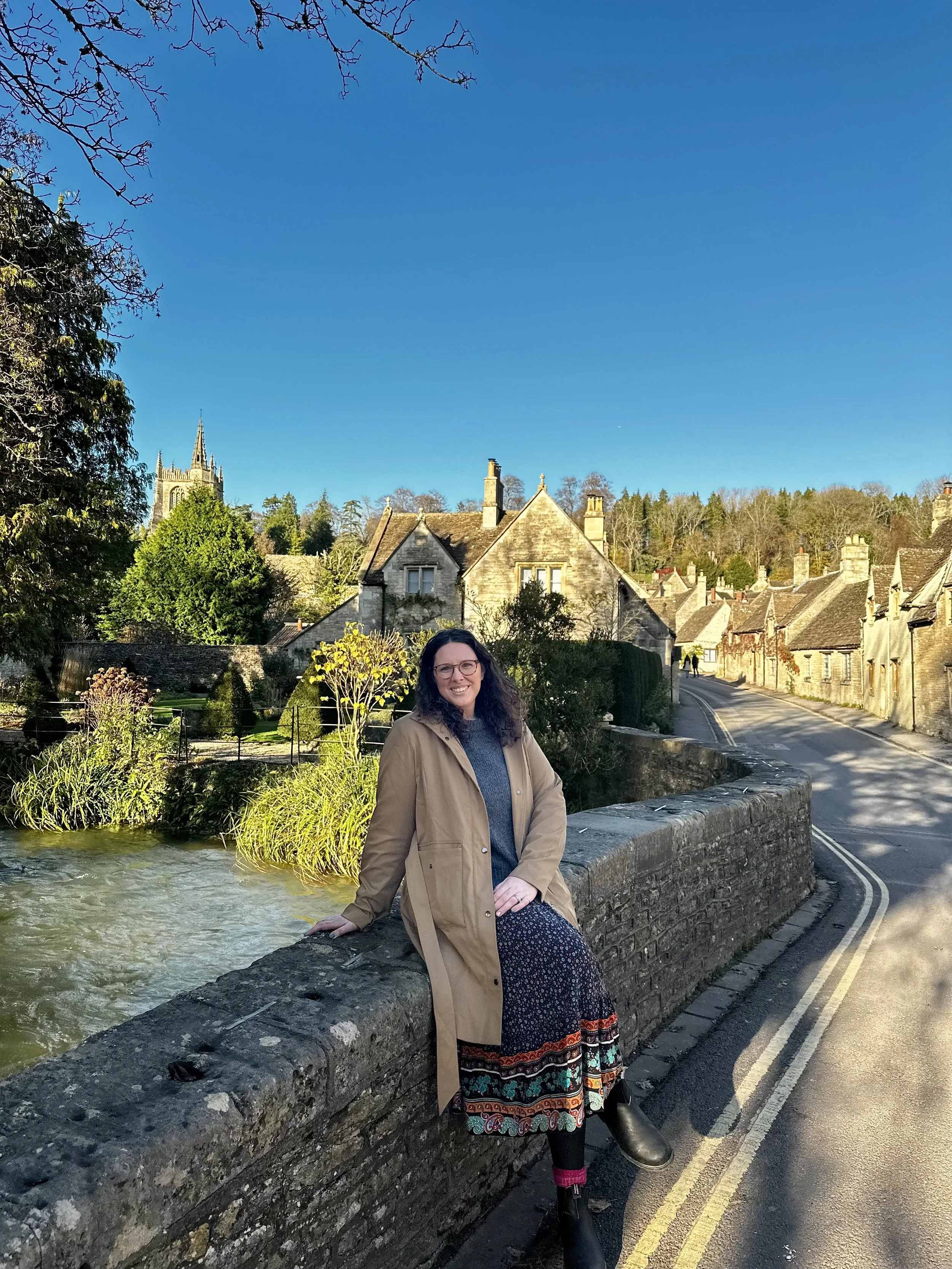 A woman with curly dark hair and glasses sitting on a stone ledge beside a small river in a quaint town. She is wearing a tan coat over a blue sweater and a patterned skirt, smiling at the camera. In the background, there are old stone houses lining a narrow road, and a church with a tall spire visible above the rooftops. The scene is set during daylight with clear blue skies and lush green trees.