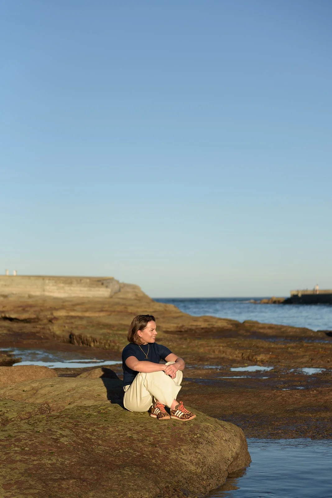 A woman sitting on a large rock by the ocean, with rocky shoreline and lighthouse visible in the background, under a clear blue sky.