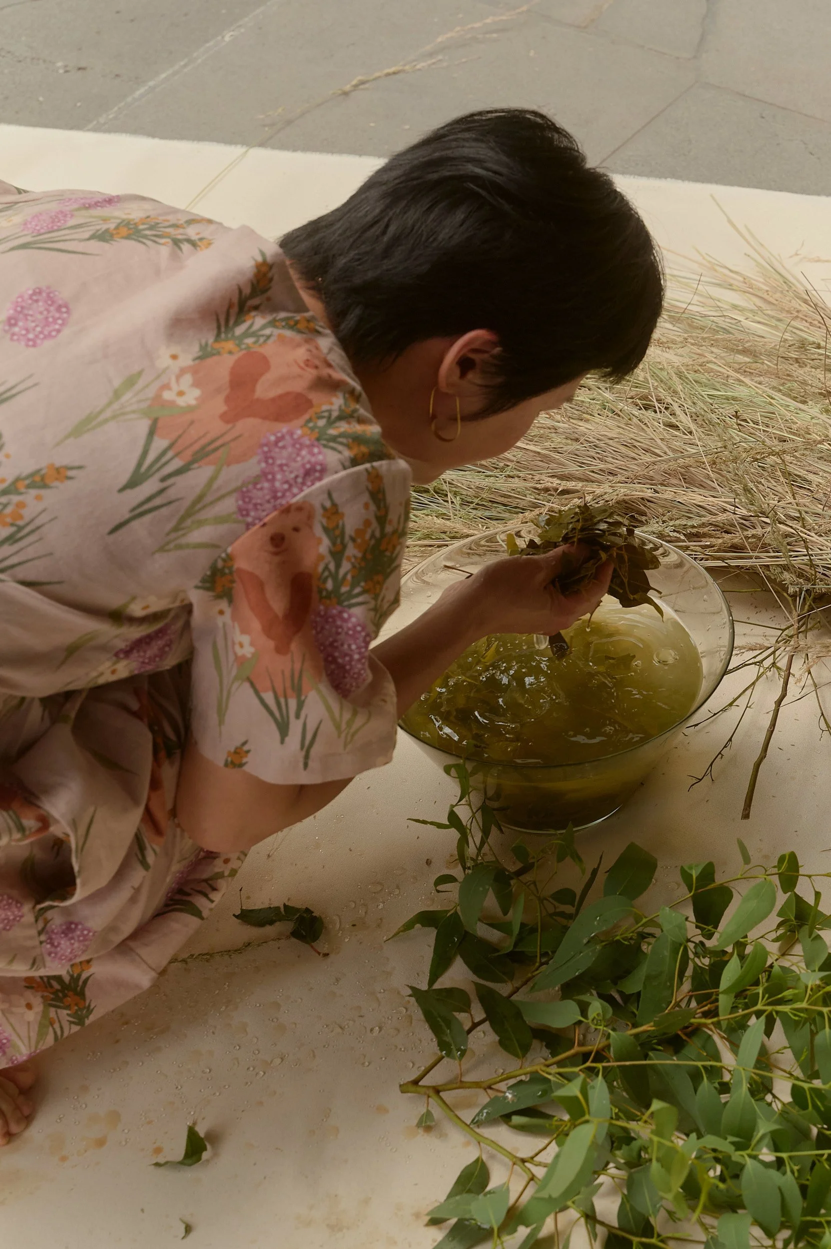 A person crouched weaving dried grasses beside a green bowl, surrounded by plants