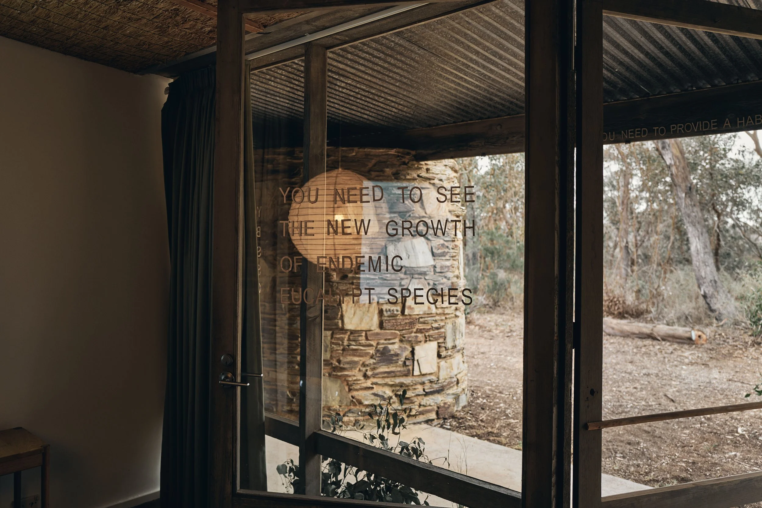 View from inside through glass lettered "You need to see the new growth of endemic eucalypt species", looking out to a stone chimney and bushland
